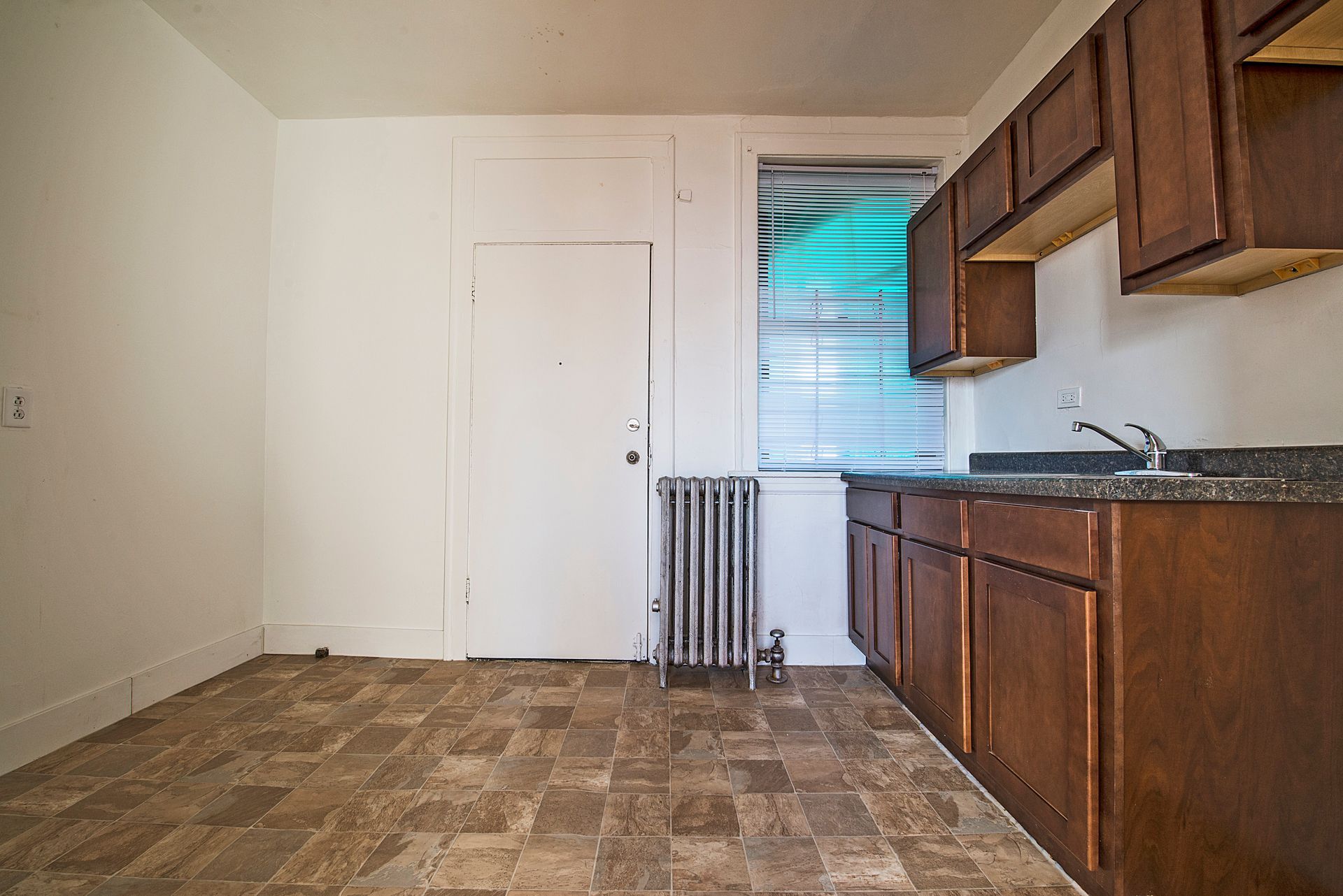 Kitchen with brown cabinets, window, door, radiator, and brown and beige tiled floor.