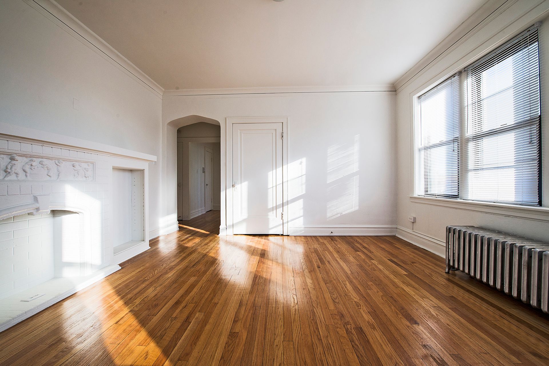 Empty room with hardwood floors, white walls, ornate fireplace, and sunlight streaming through a window.