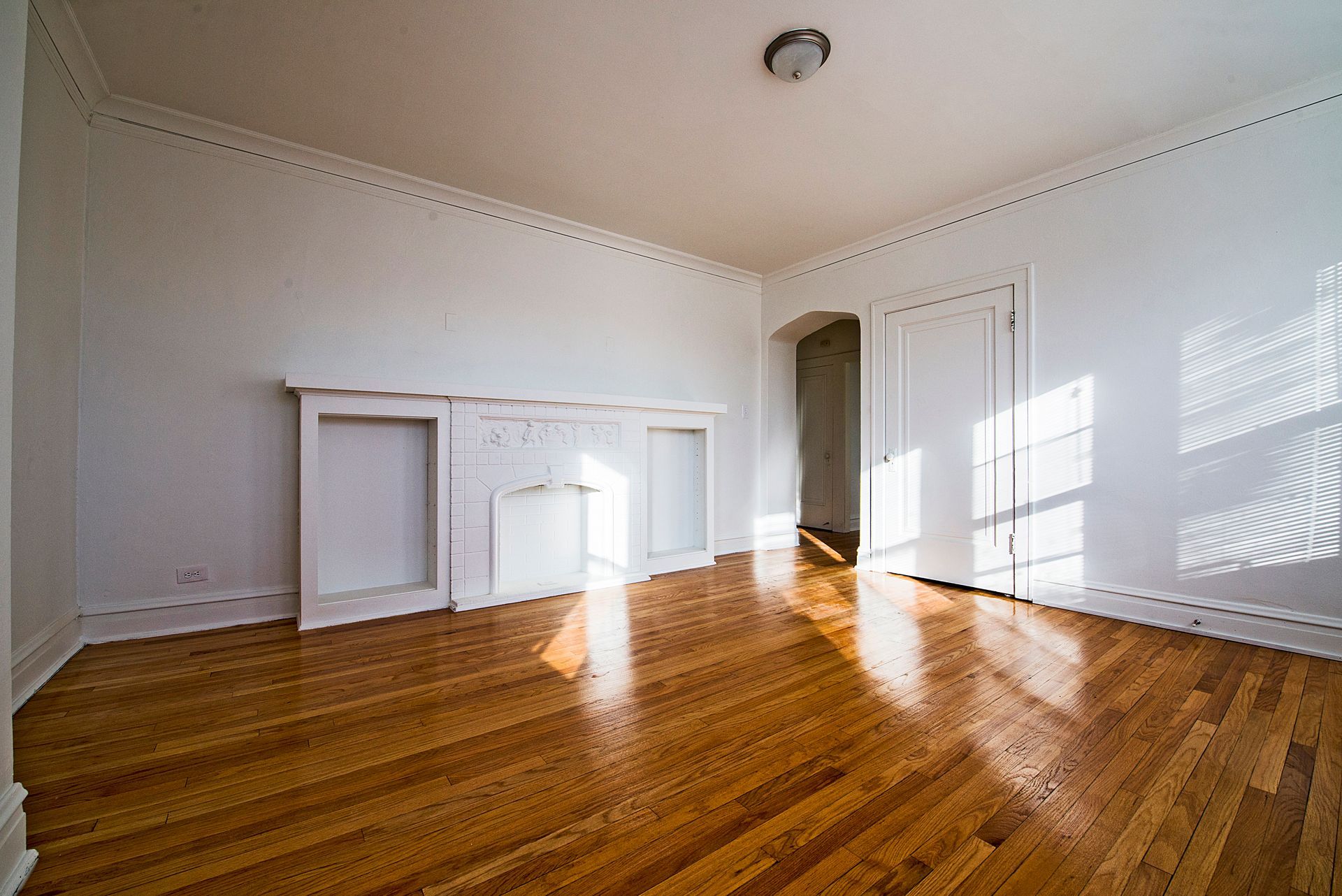 Empty room with hardwood floors, white walls, and a built-in fireplace. Sunlight streams through a window.