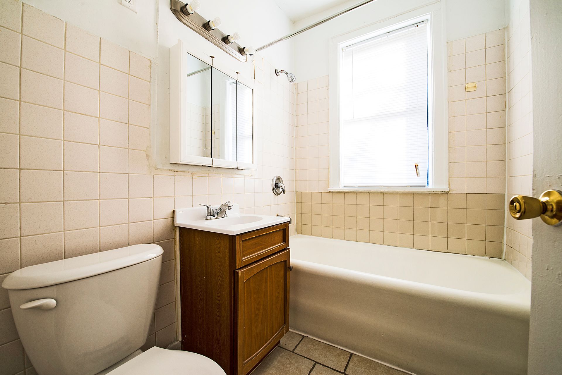 Small bathroom with toilet, sink, bathtub, and window with closed blinds. Cream-colored tile walls.