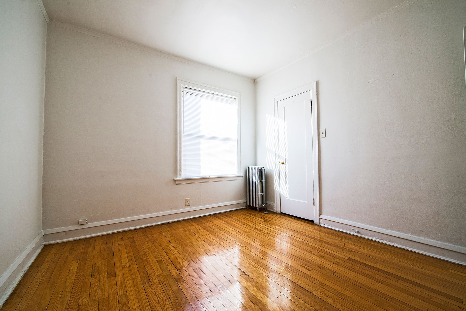 Empty room with hardwood floors, a window with blinds, and a closed door.