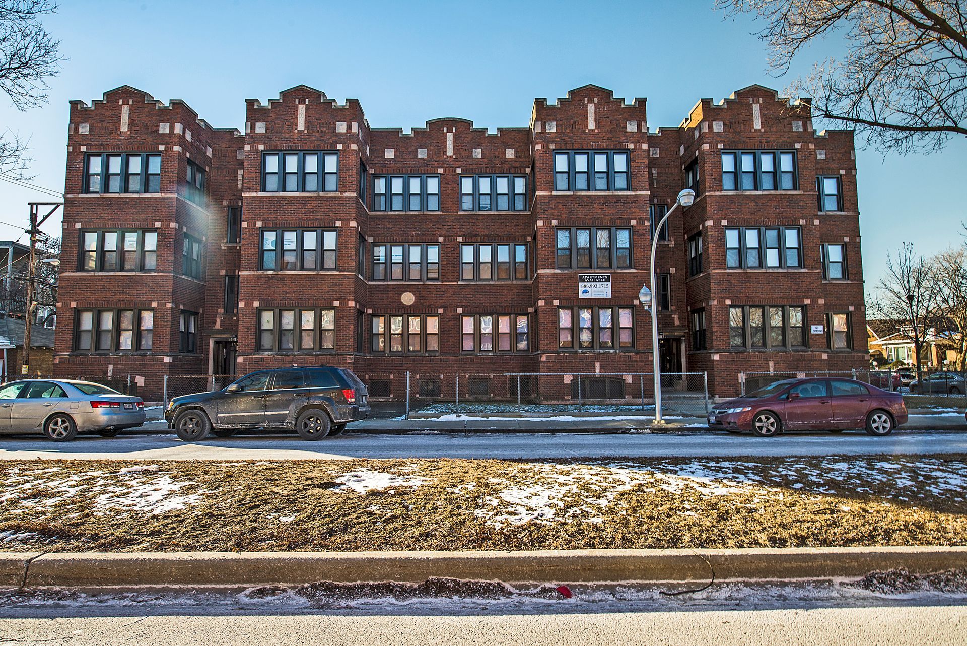 Brown brick apartment building with cars parked out front, snow on the ground, sunny day.
