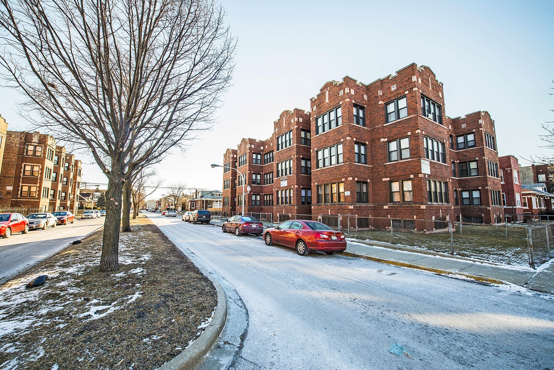 Red brick apartment buildings line a snow-covered street with parked cars and a bare tree.