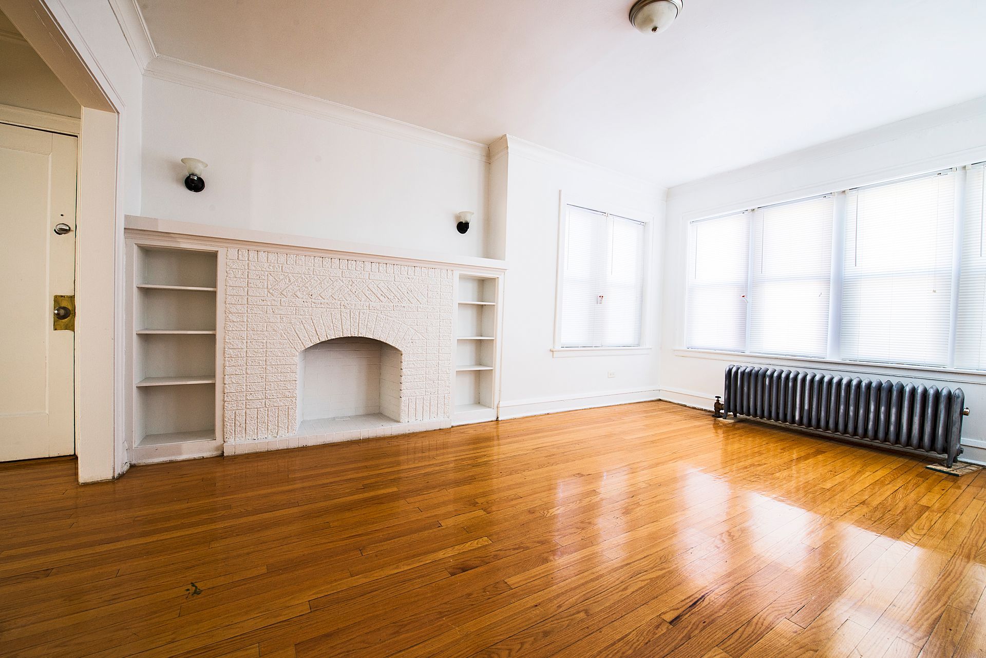 Empty living room with hardwood floors, white brick fireplace, and large windows.