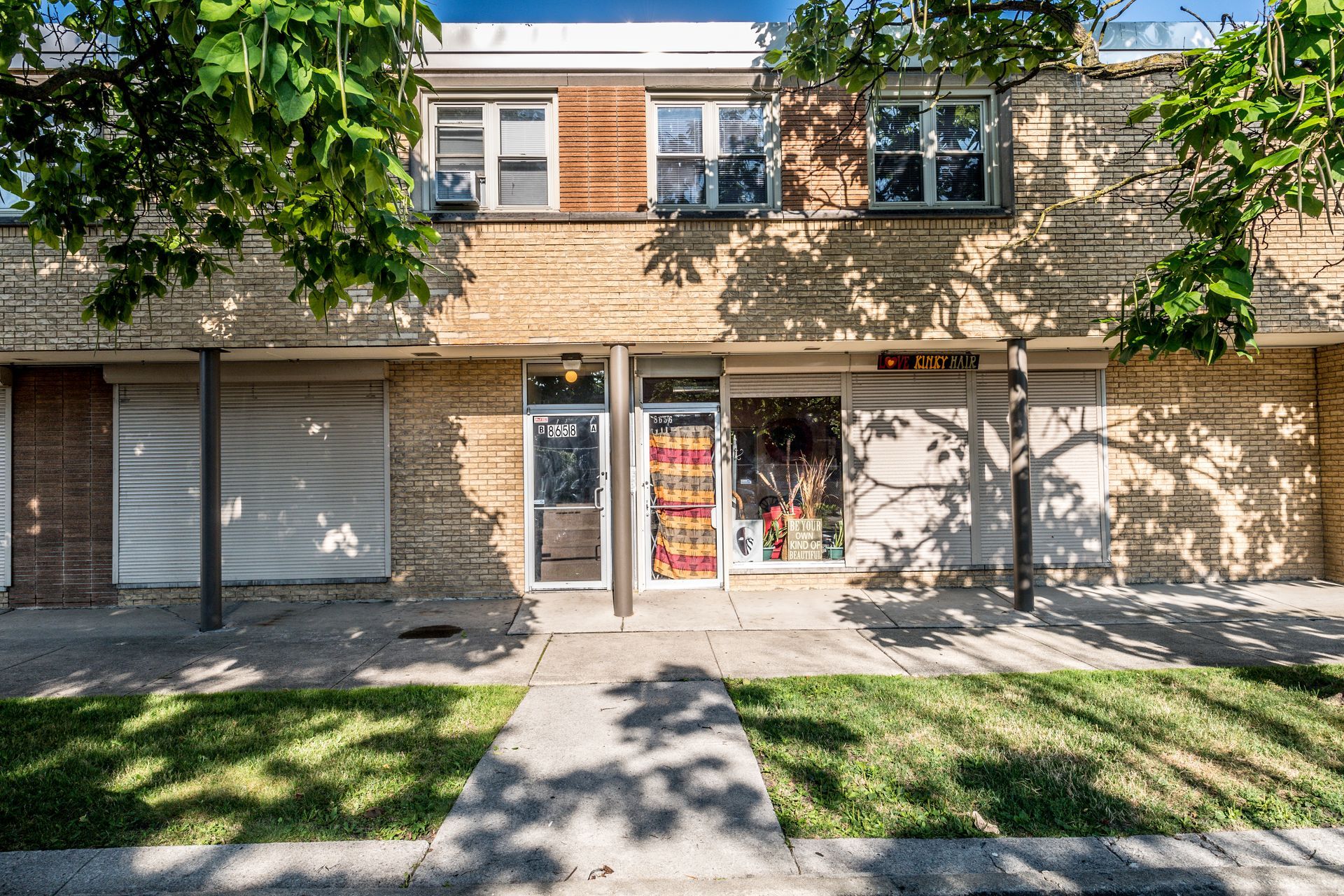 Two-story brick building with storefronts and a sidewalk in front. Trees cast shadows.