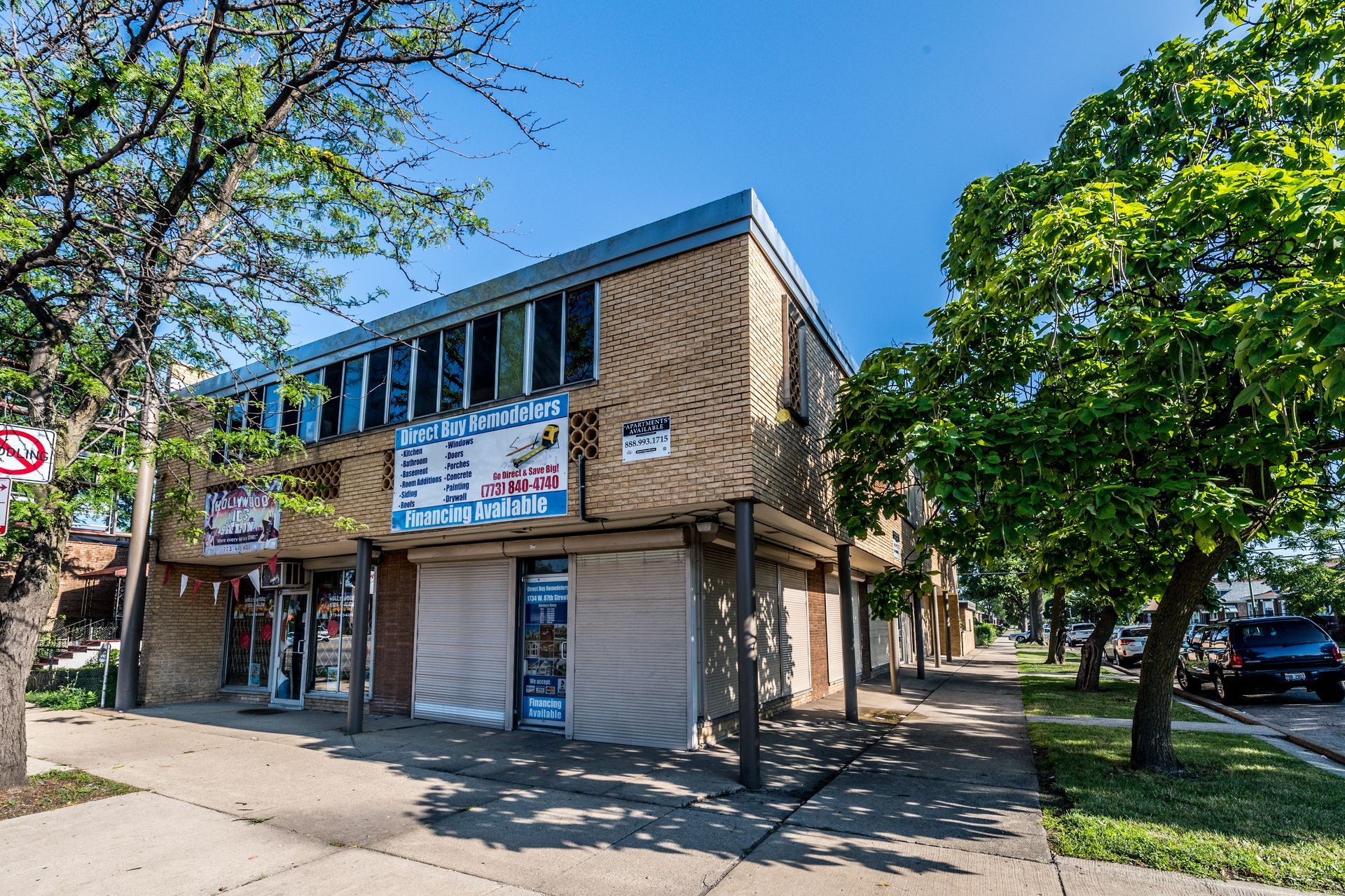 Two-story commercial building on a city street; closed storefronts, brick facade, trees, sunny day.