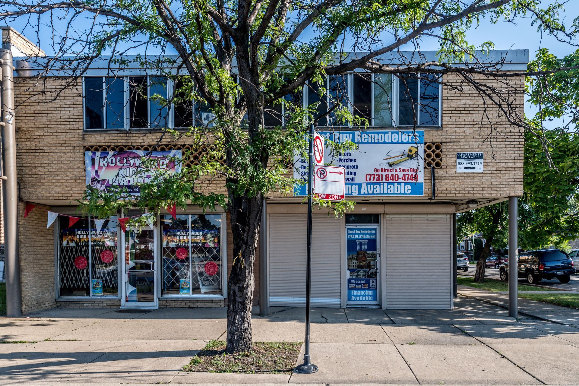Two-story brick building with businesses on the ground floor, a tree in front, and a blue sky.