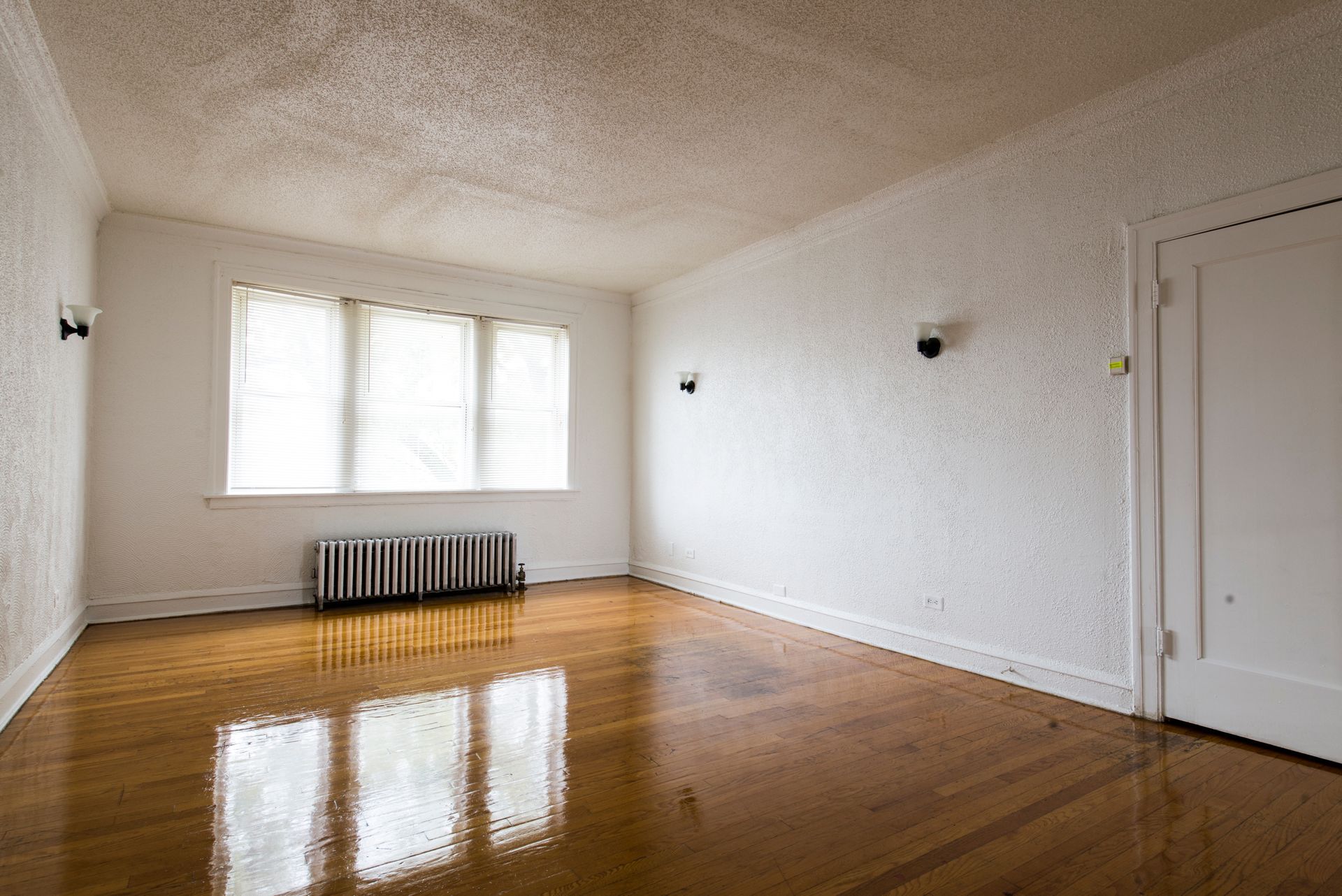 Empty room with hardwood floors, textured white walls, window with curtains, radiator, and a closed door.