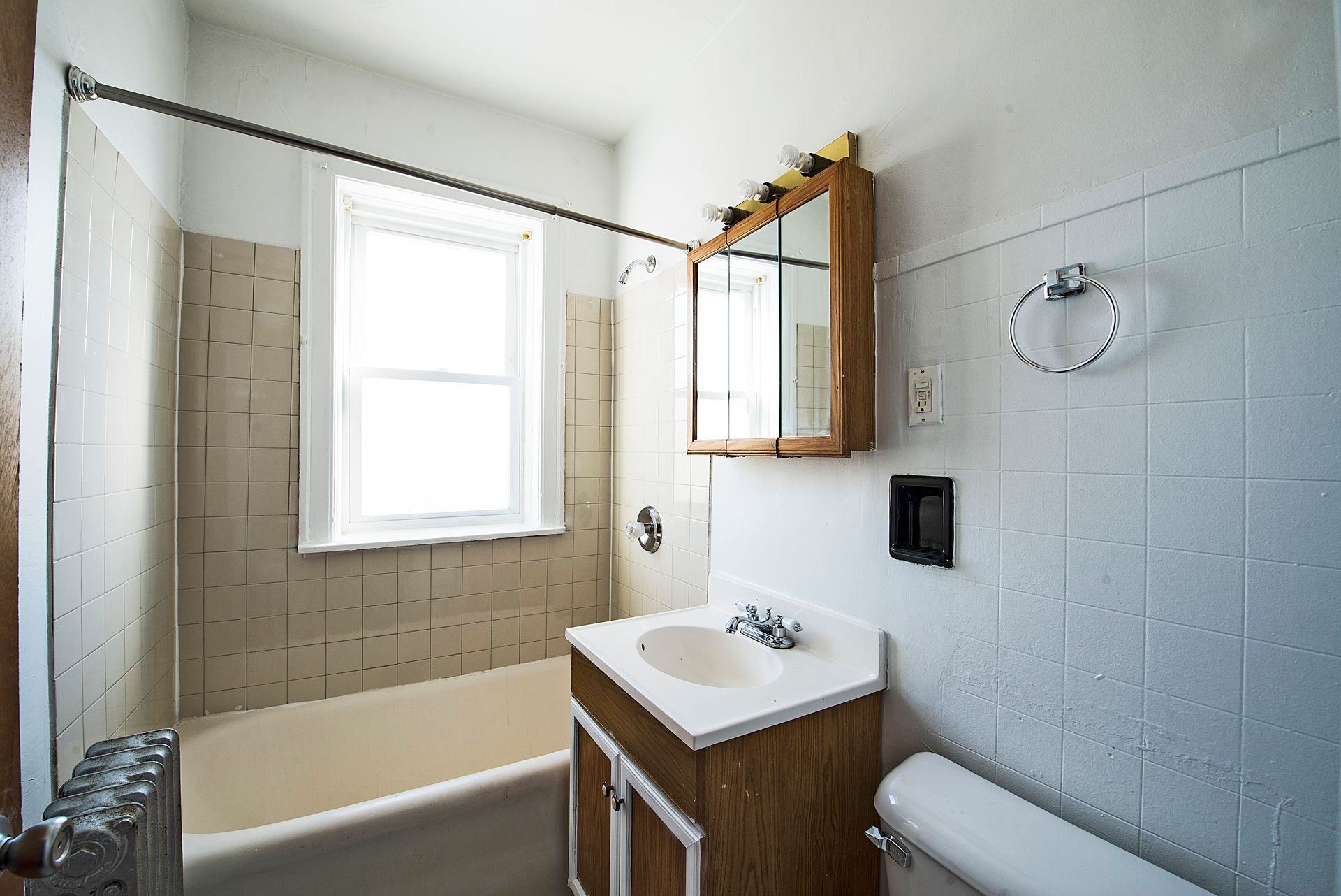 Bathroom with bathtub, sink, mirror, and toilet. Beige and white tiled walls, window, and wood vanity.