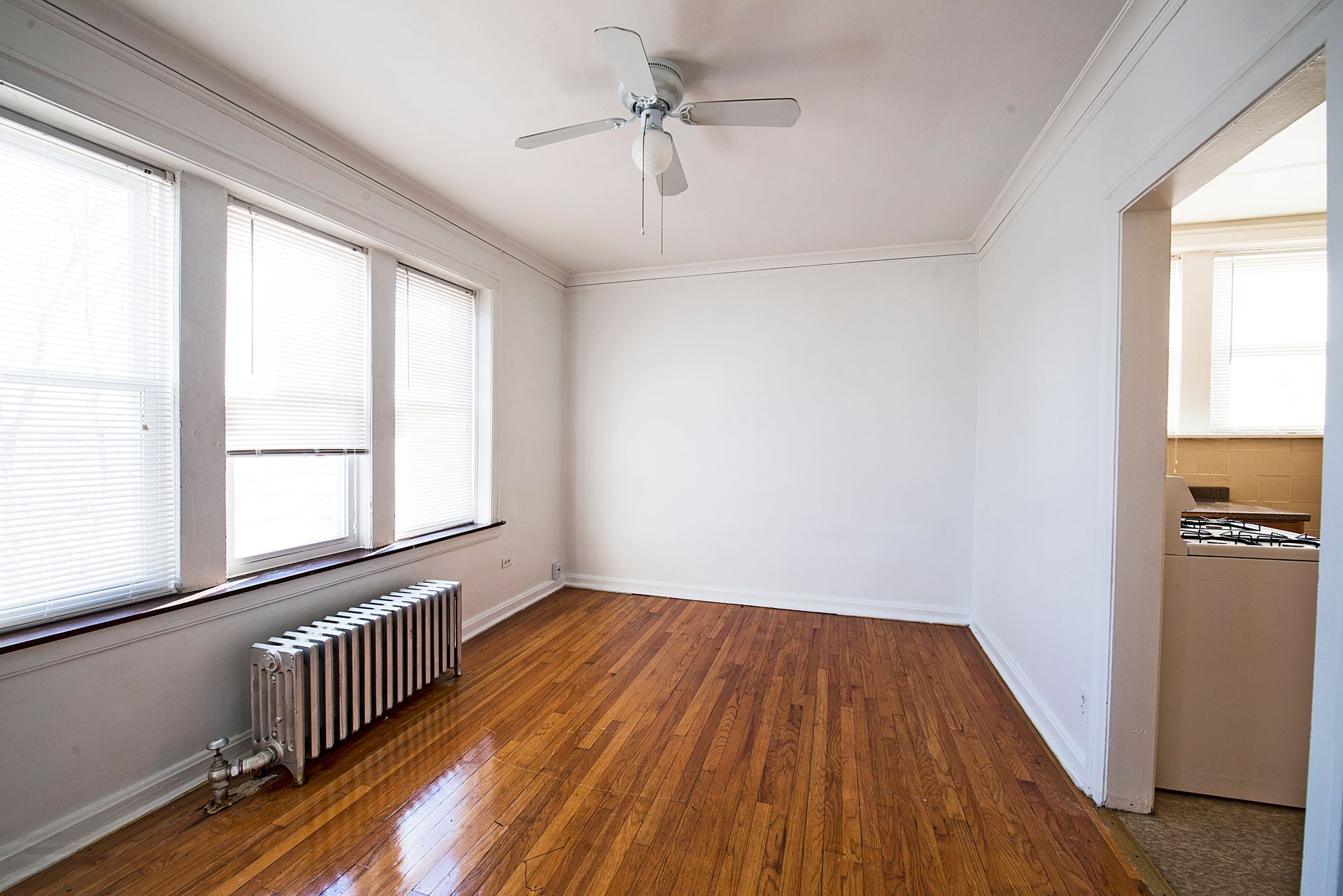 Empty room with wood floors, blinds, radiator, ceiling fan, and doorway to a kitchen.