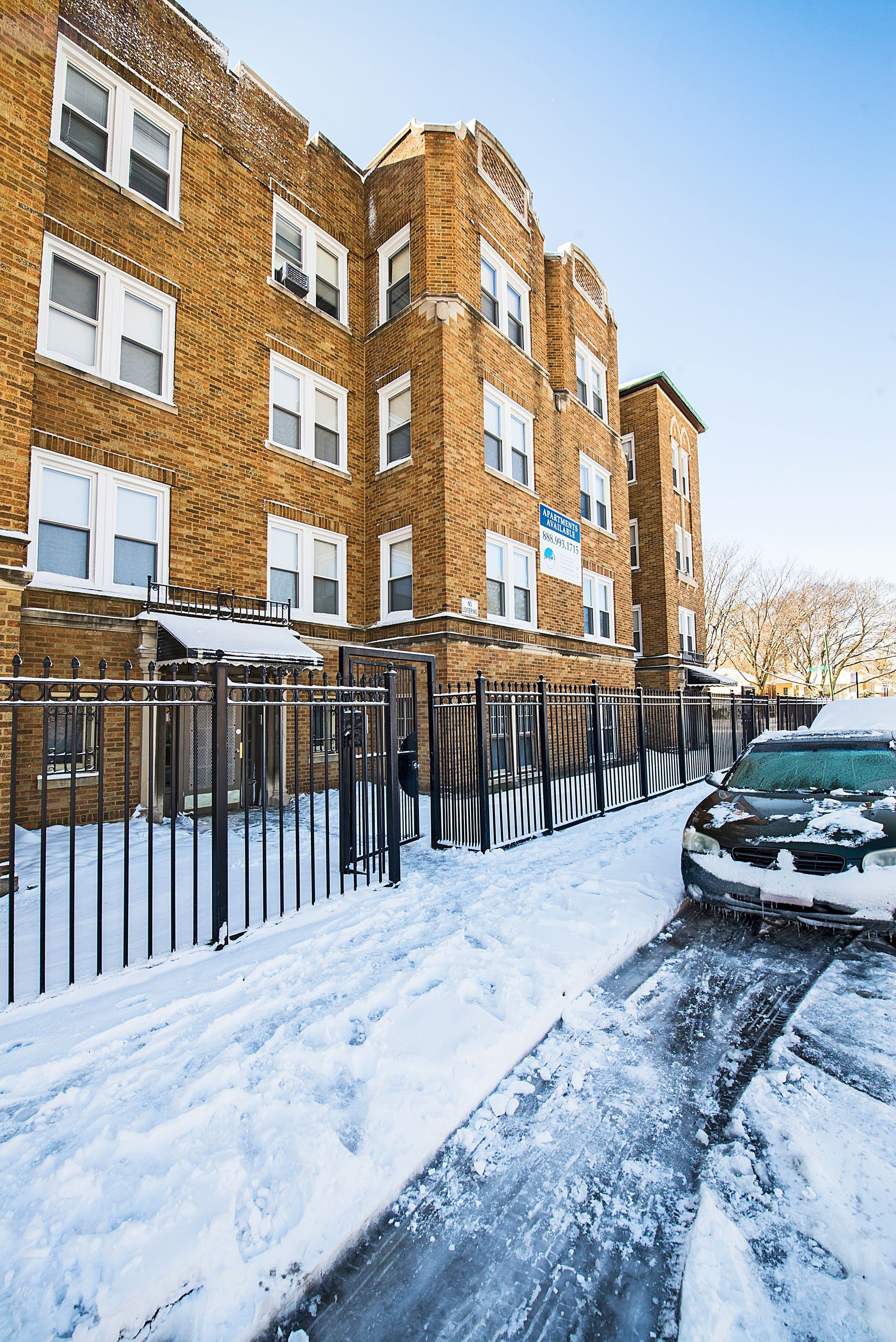 Snow-covered brick apartment building with a fence and a car covered in frost on a snowy street.