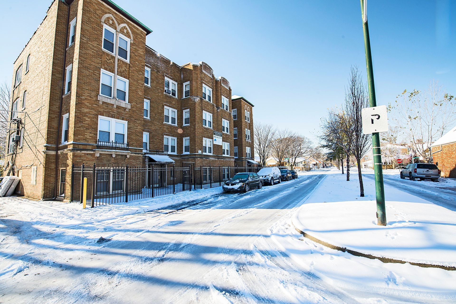 Snowy street scene with parked cars, a brick apartment building, and a parking sign.