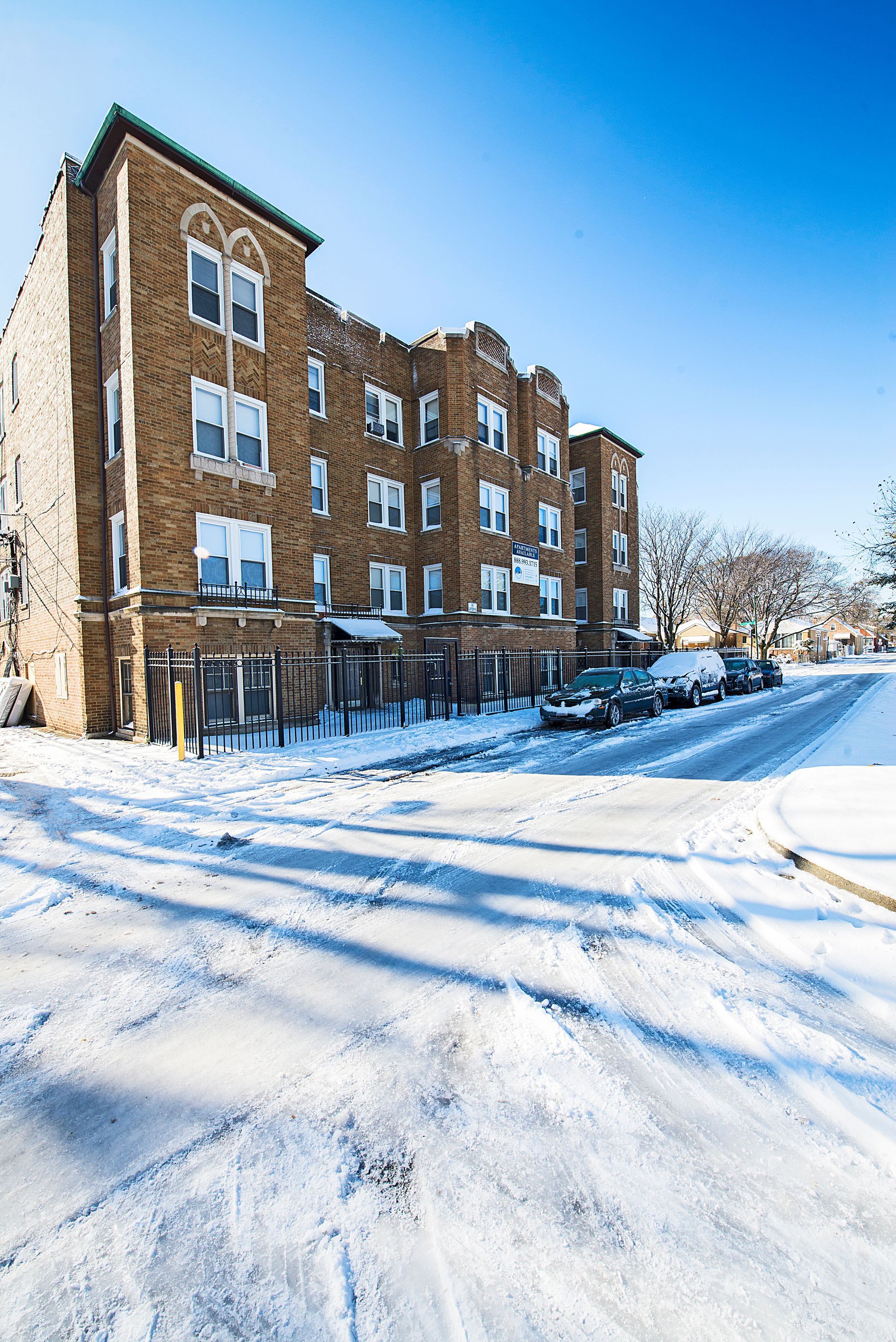 Snowy street with multi-story brick apartment building. Cars parked along the curb. Blue sky.