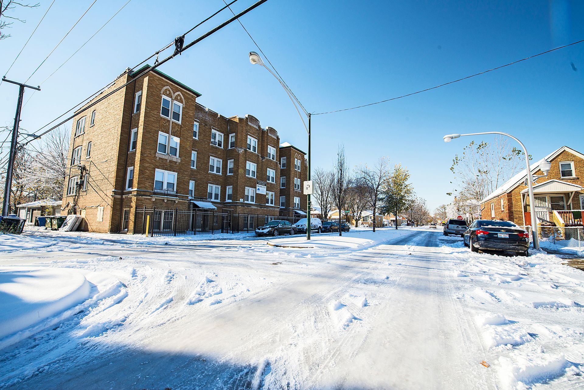 Snow-covered street in front of a brick apartment building and houses; bright sunny day.