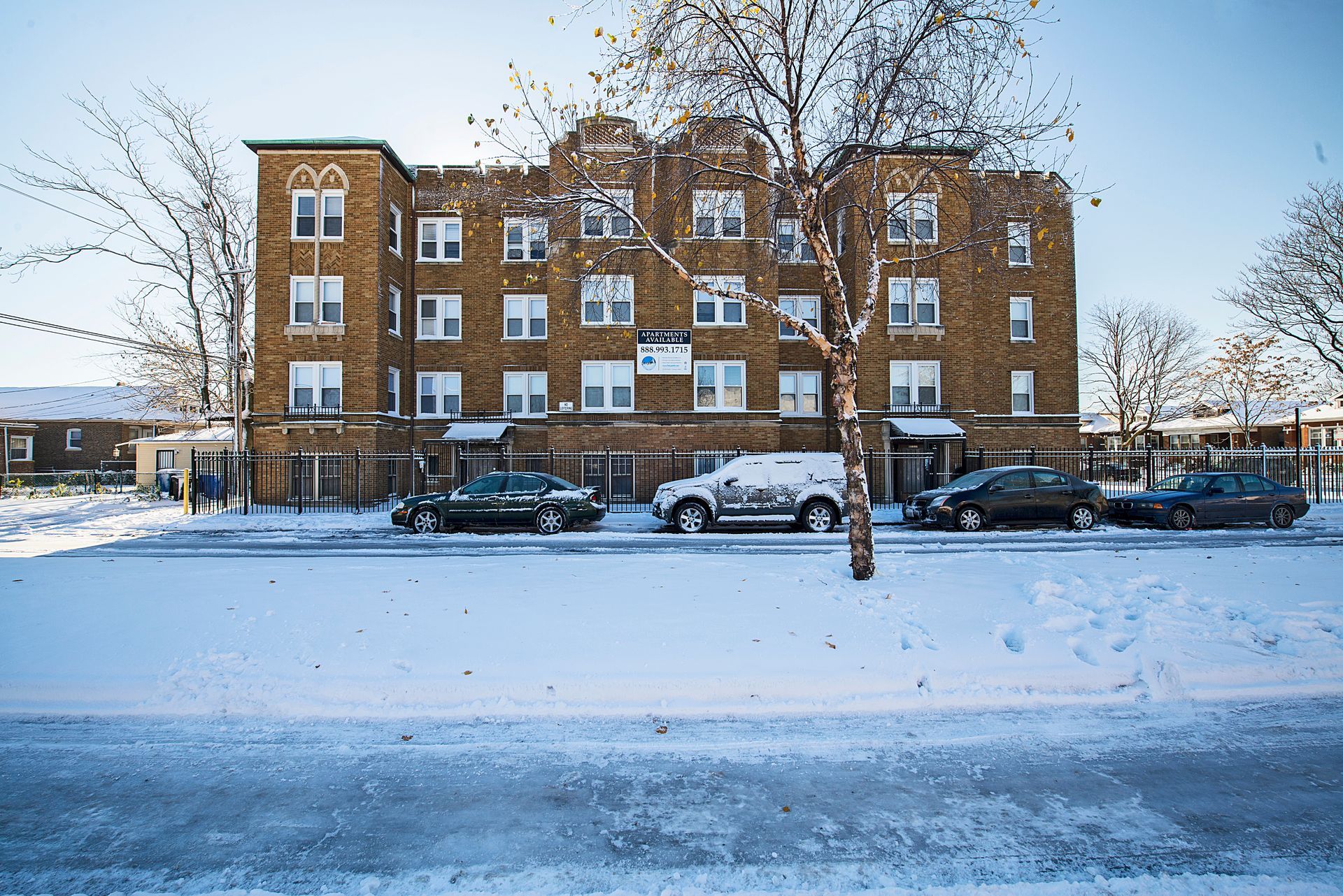 Snow-covered street with a brick apartment building and parked cars on a clear day.