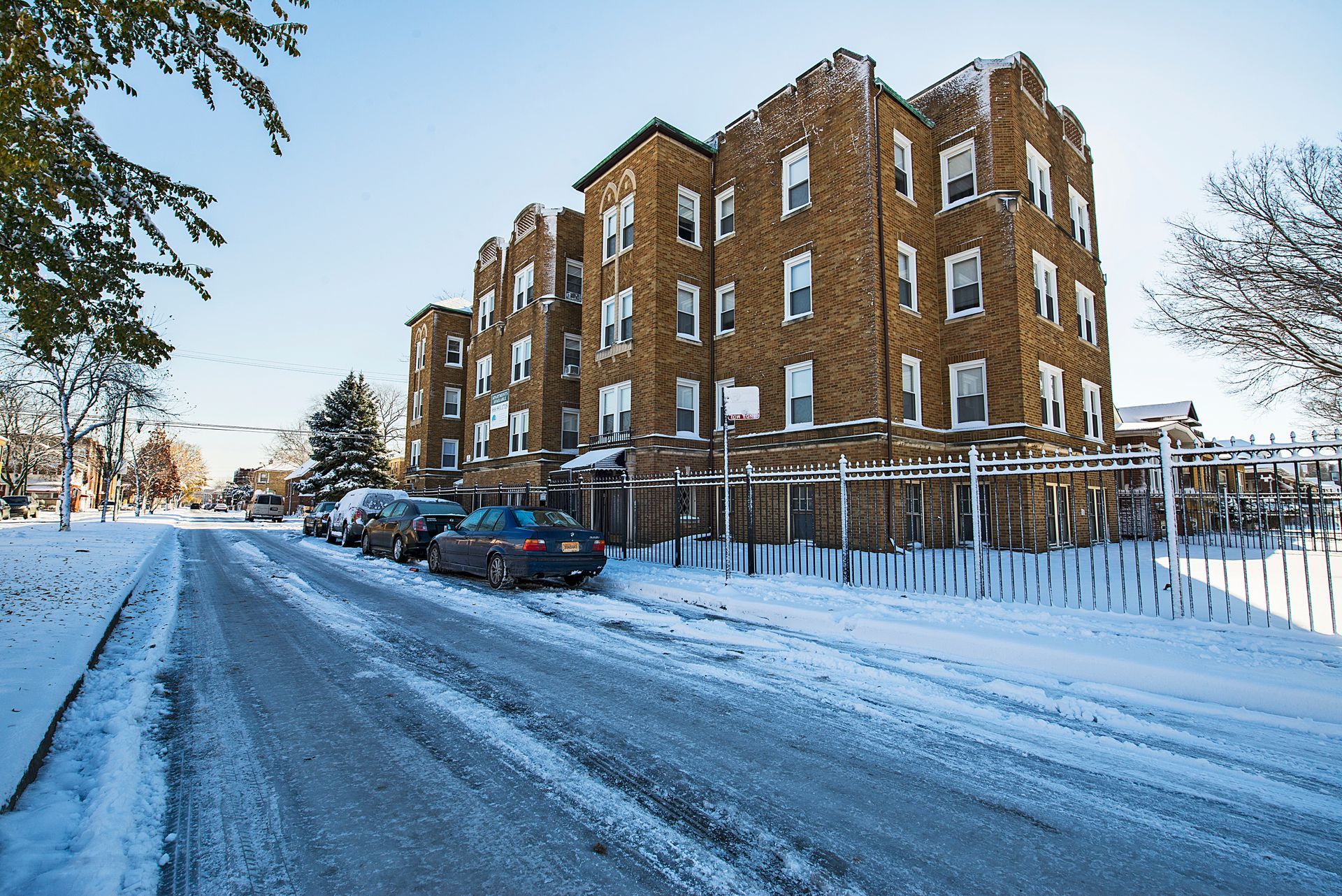 A multi-story brick apartment building on a snow-covered street with parked cars. Blue sky.