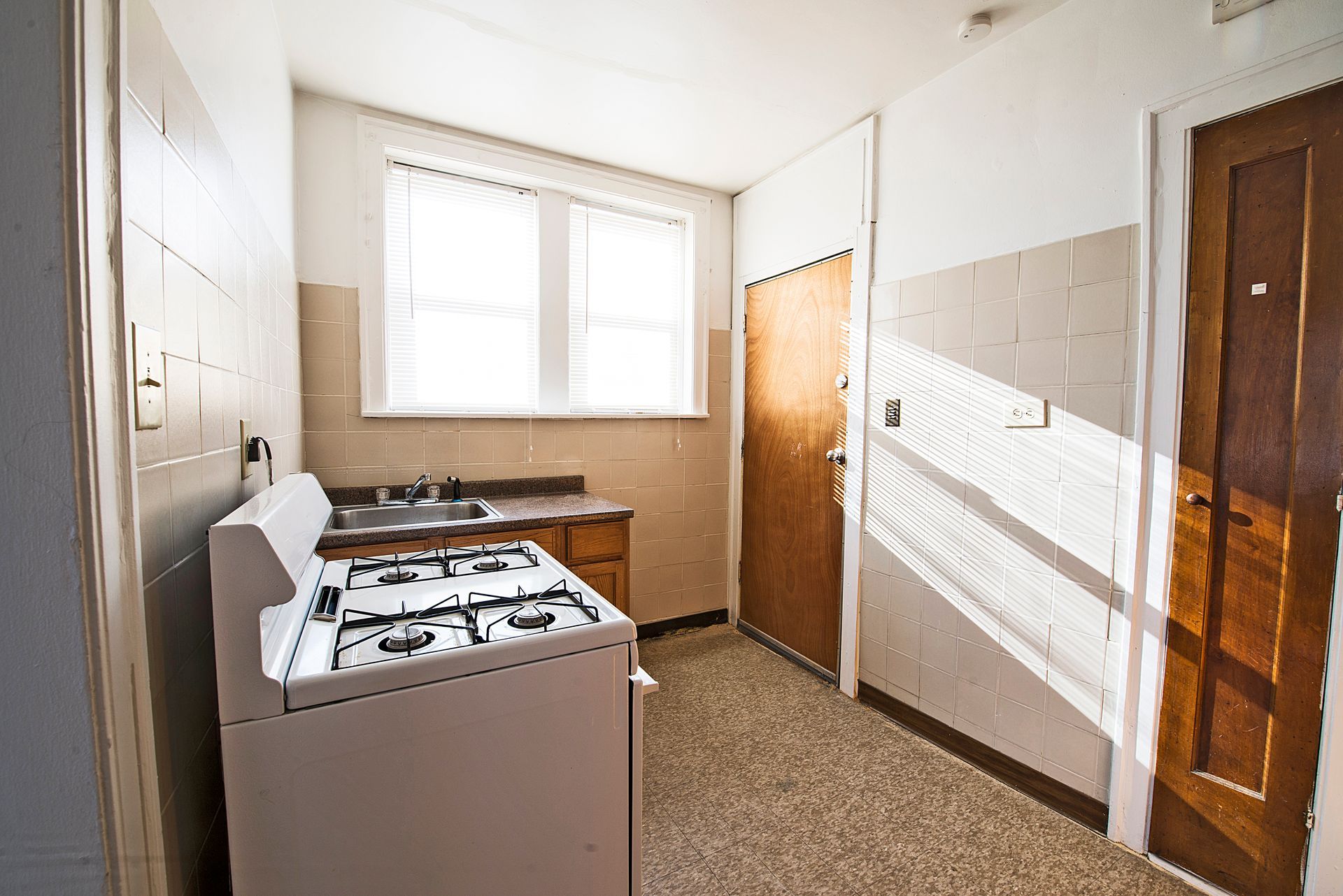 Small kitchen with white stove, sink, and wooden cabinets. Window provides natural light.