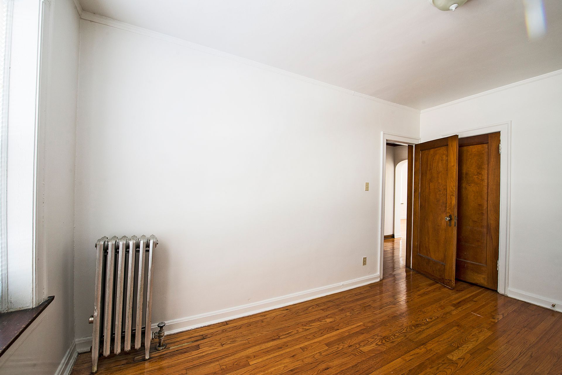 Empty room with white walls, hardwood floor, radiator, and two wood doors.