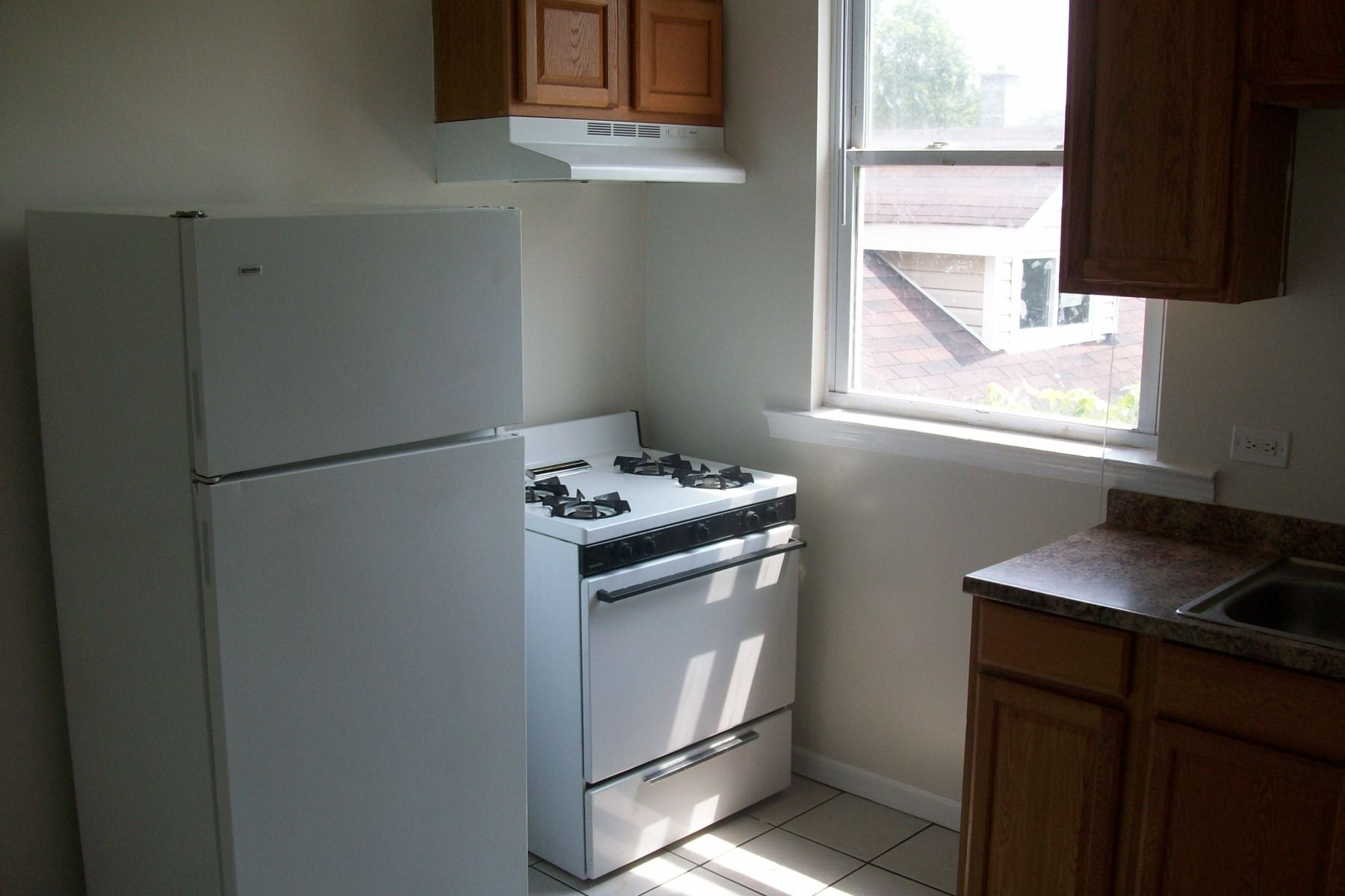 A small kitchen with white appliances, wooden cabinets, and a window.