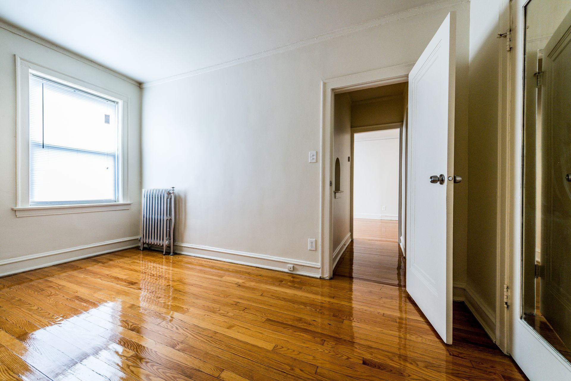 Empty room with hardwood floors, a window, radiator, and open door to another room.