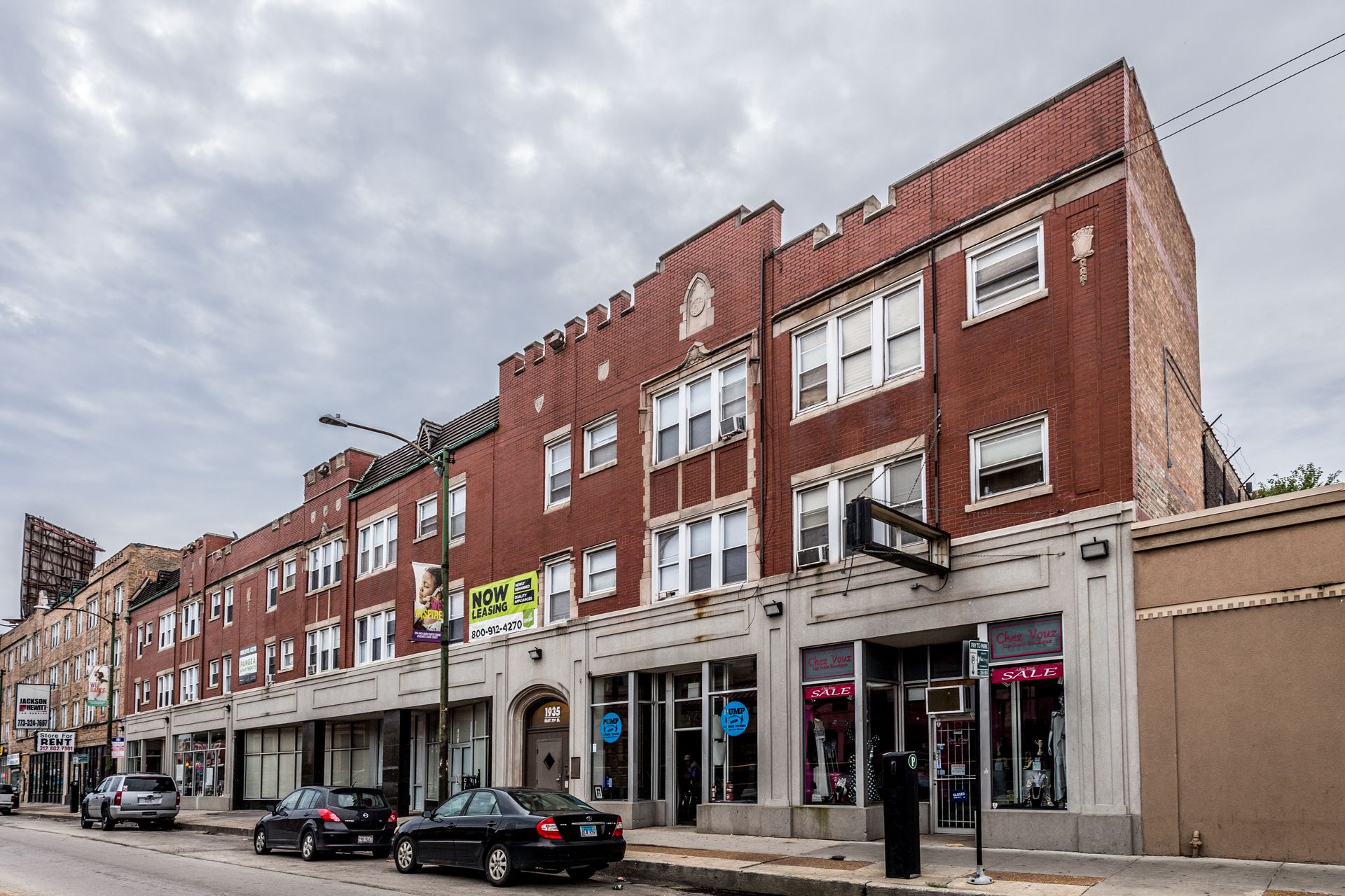Brick building with storefronts. Dark cars parked in front. Overcast sky.