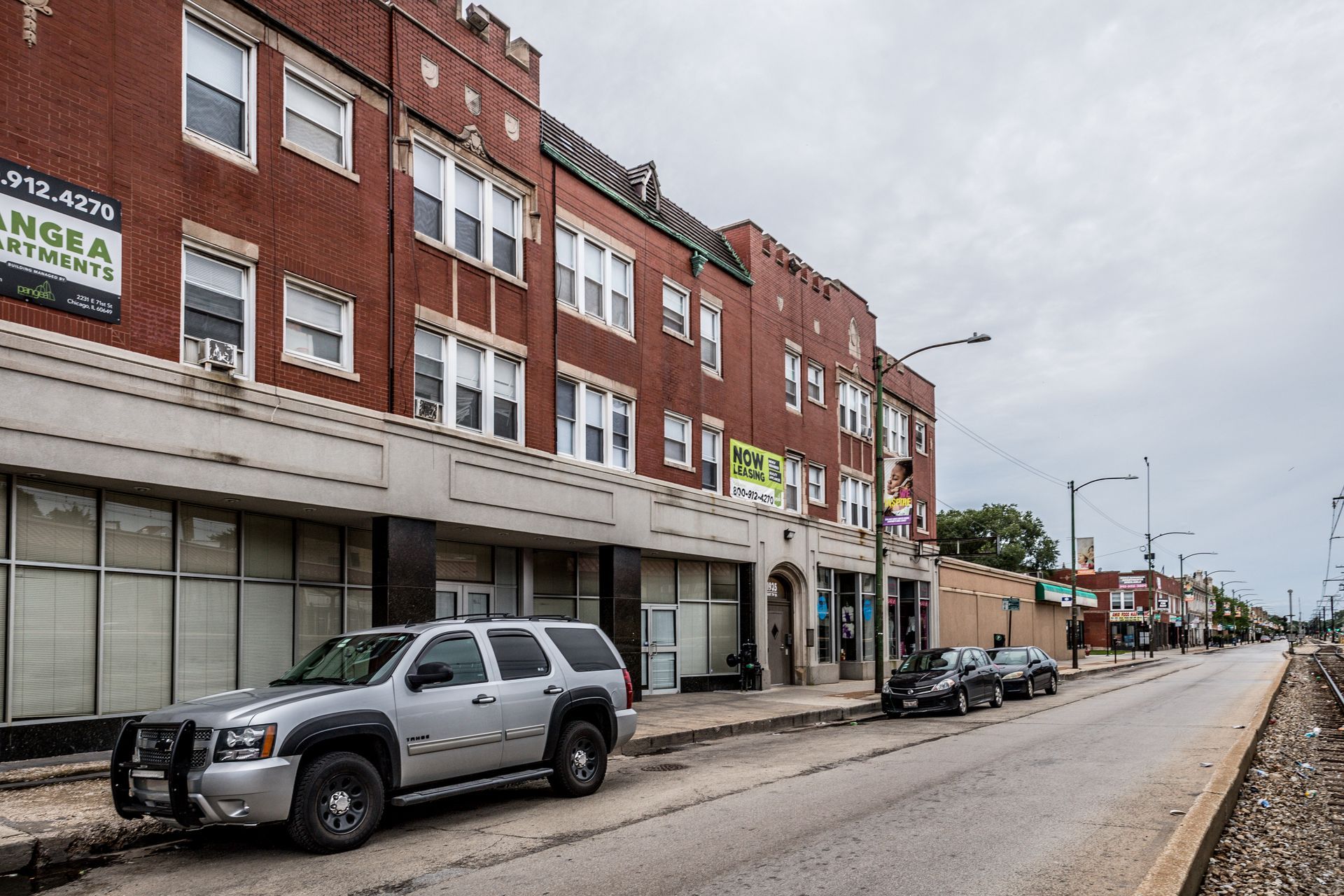 Brick apartment building with vacant storefronts, gray SUV parked on street.