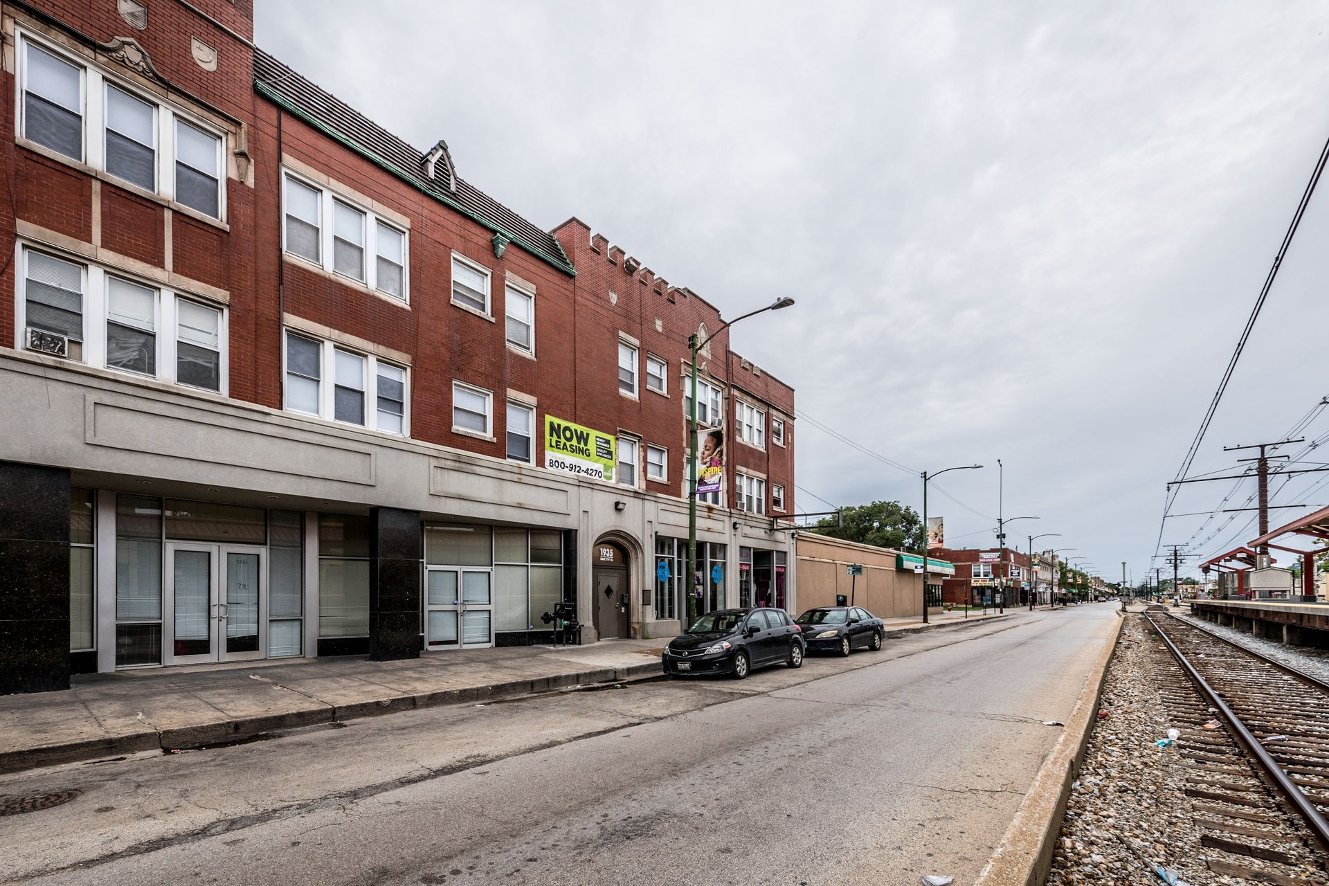 Brick building with storefronts on a street next to train tracks under a cloudy sky.