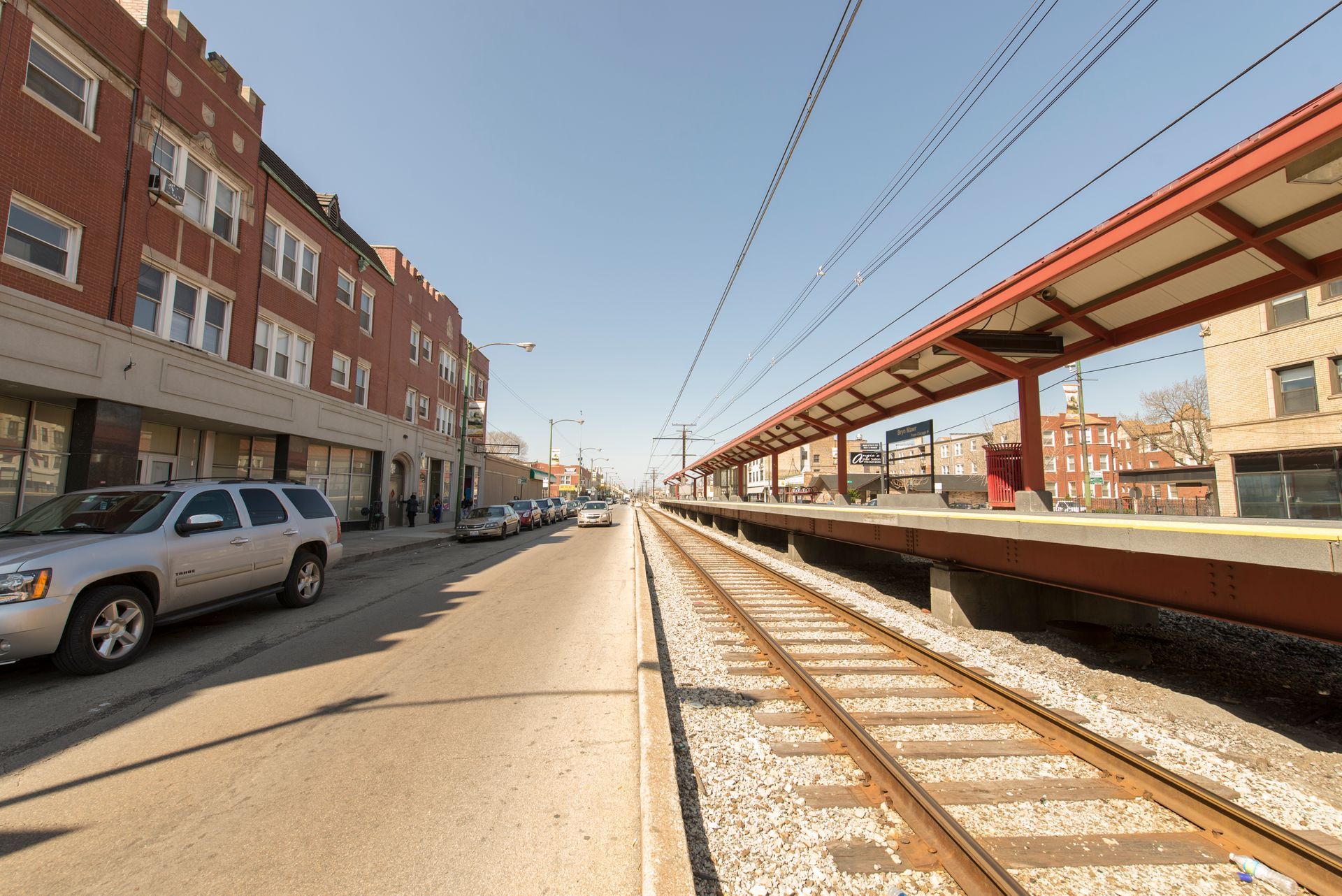 Train tracks alongside a street with parked cars and a red brick building on a sunny day.