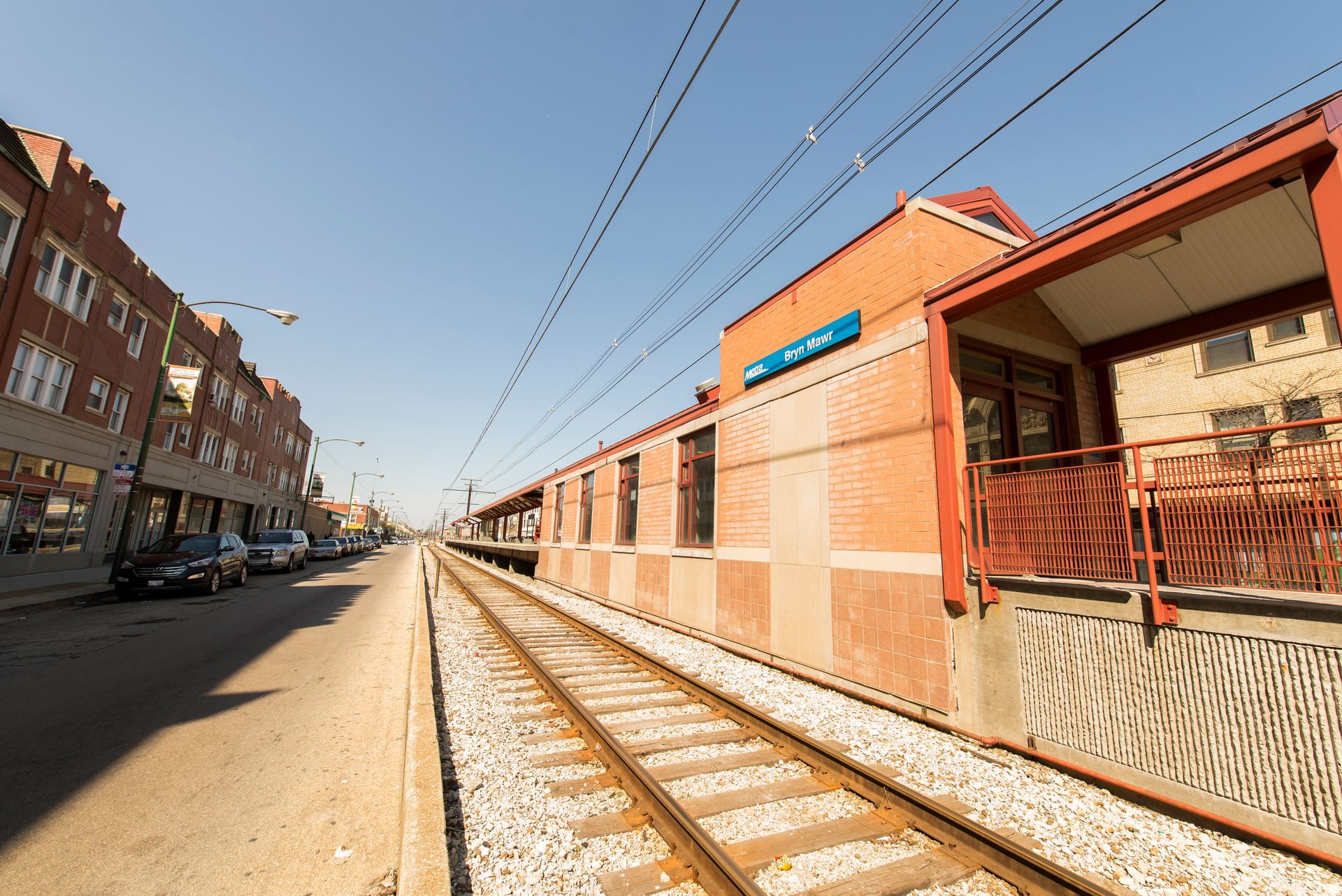 Train tracks next to a red brick station and storefronts under a clear blue sky.