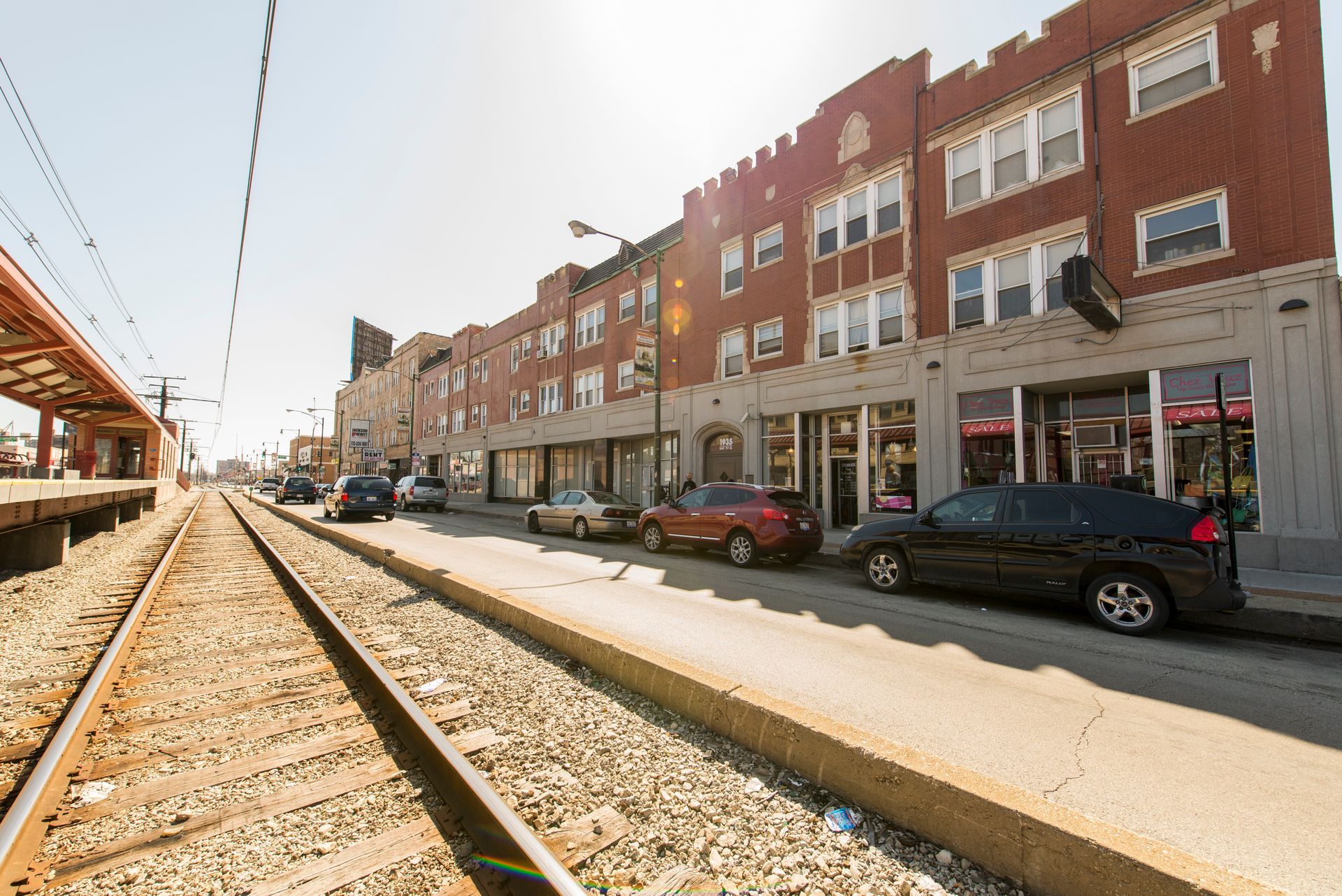 Street scene with buildings, parked cars, and train tracks. Bright sunlight.
