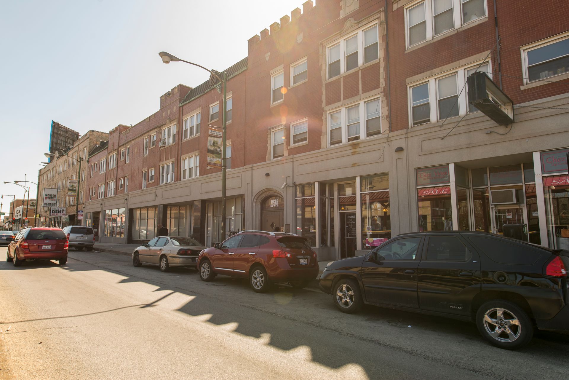 Street view of a brick building with shops on the ground floor and apartments above, cars parked along the street.