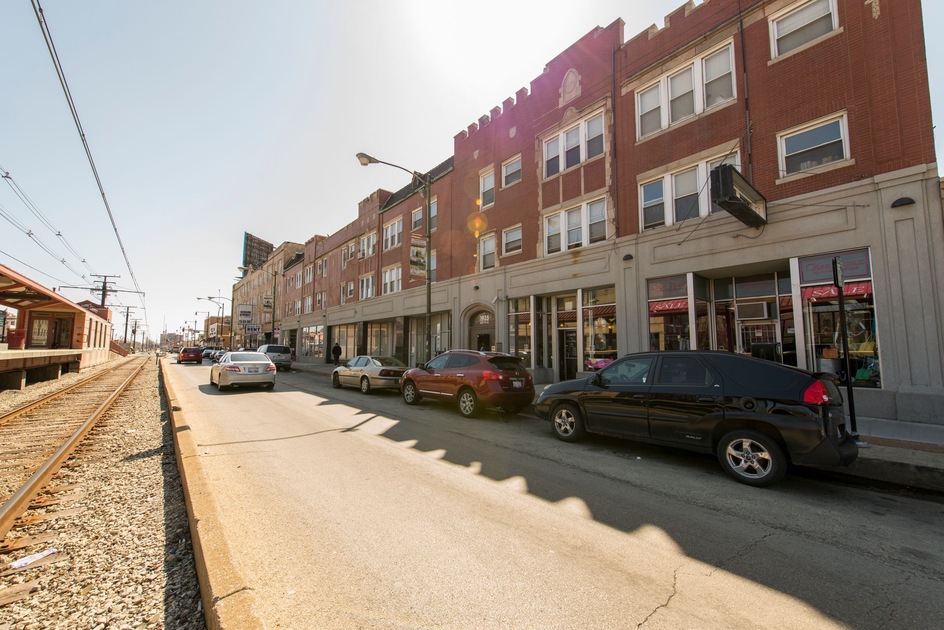 Street scene with brick buildings, parked cars, train tracks, and a sunny sky.