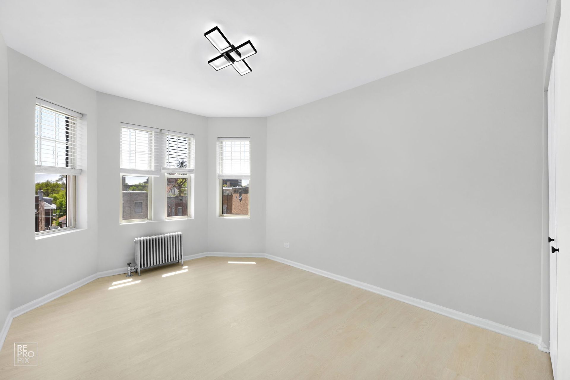 Empty room with light-colored flooring, white walls, and three windows. Includes a radiator and a modern ceiling light.