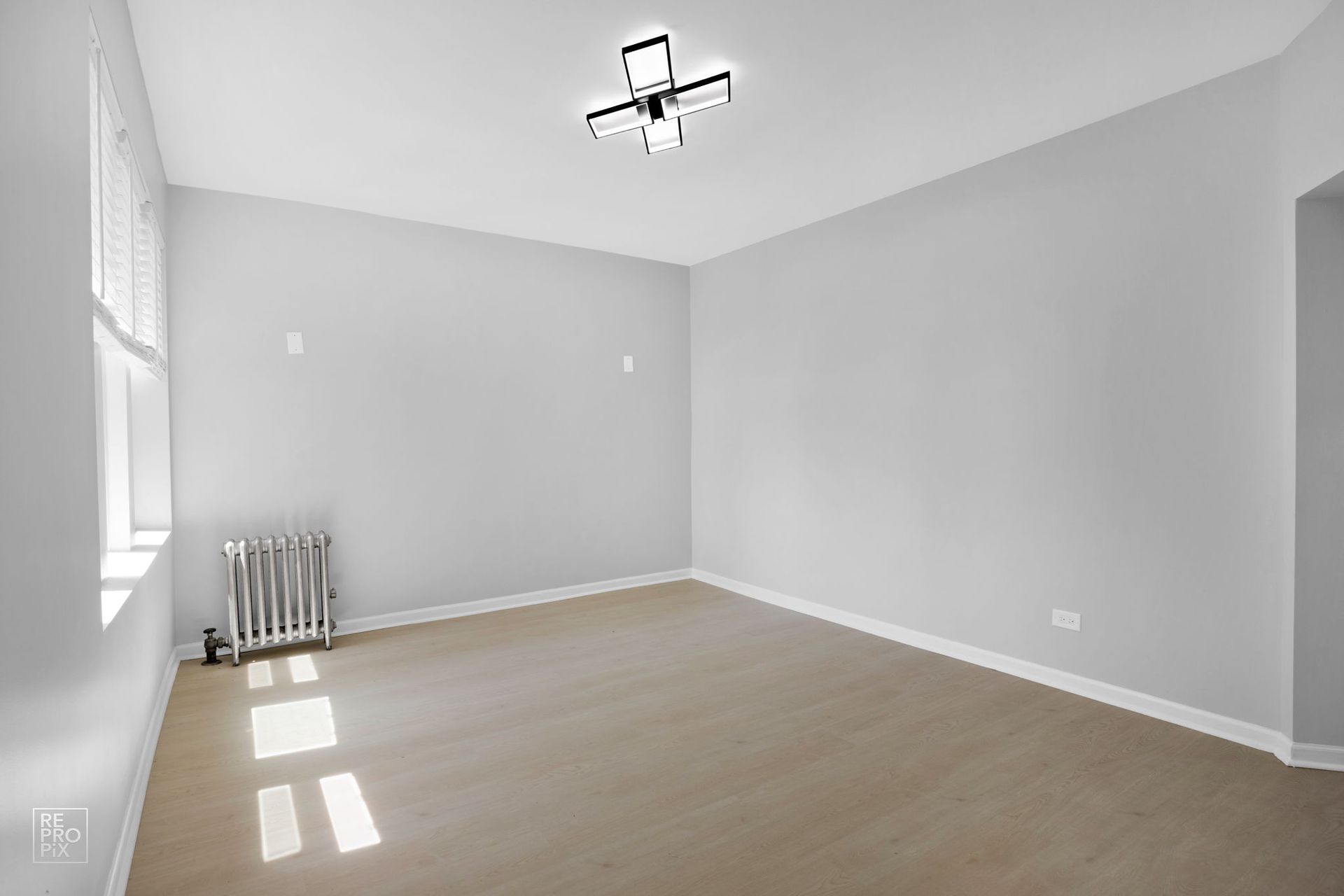 Empty room with pale grey walls, tan floor, radiator, and a light fixture.