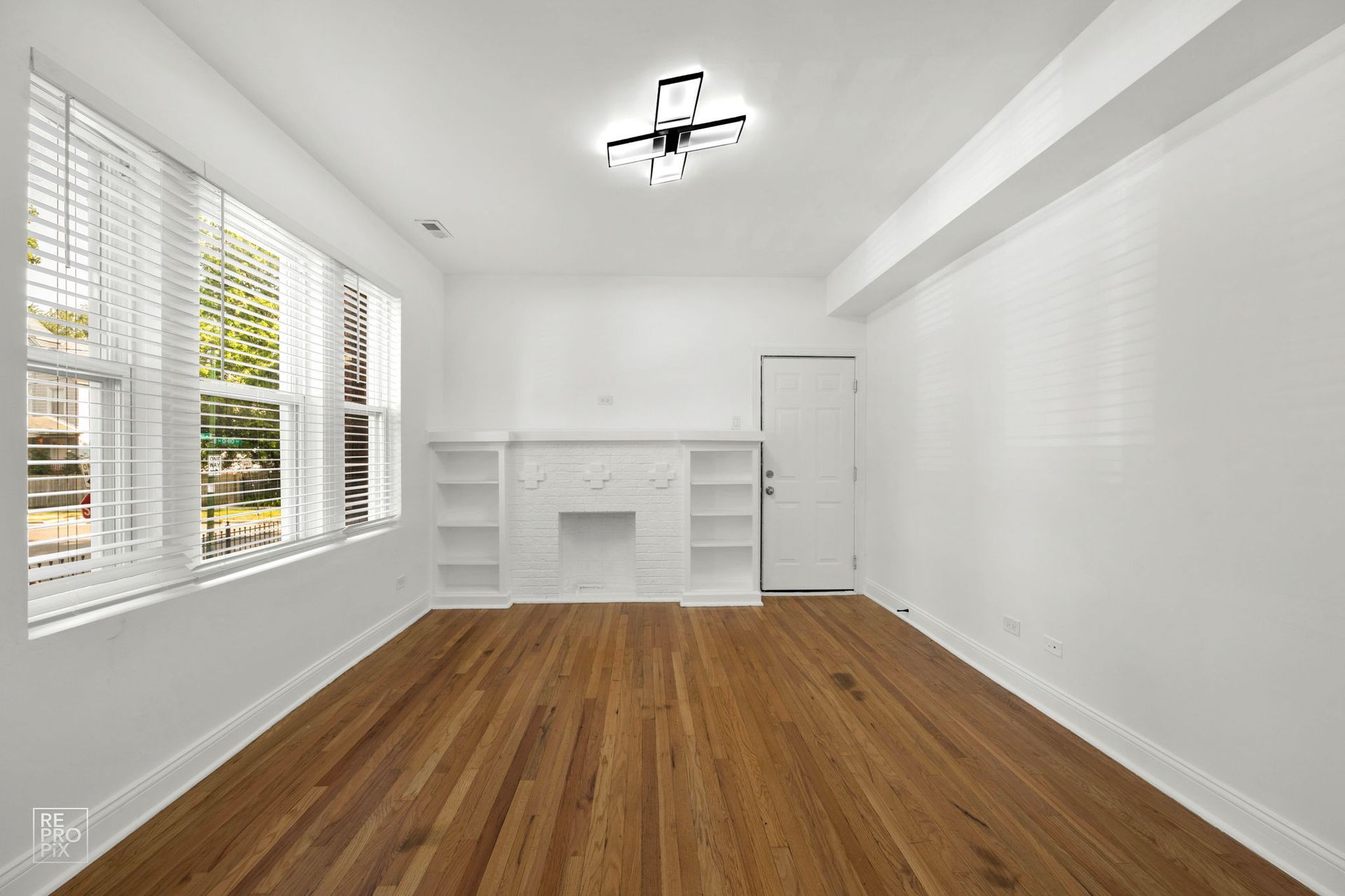 Empty room with hardwood floors, white walls, built-in shelves, and a window with blinds.