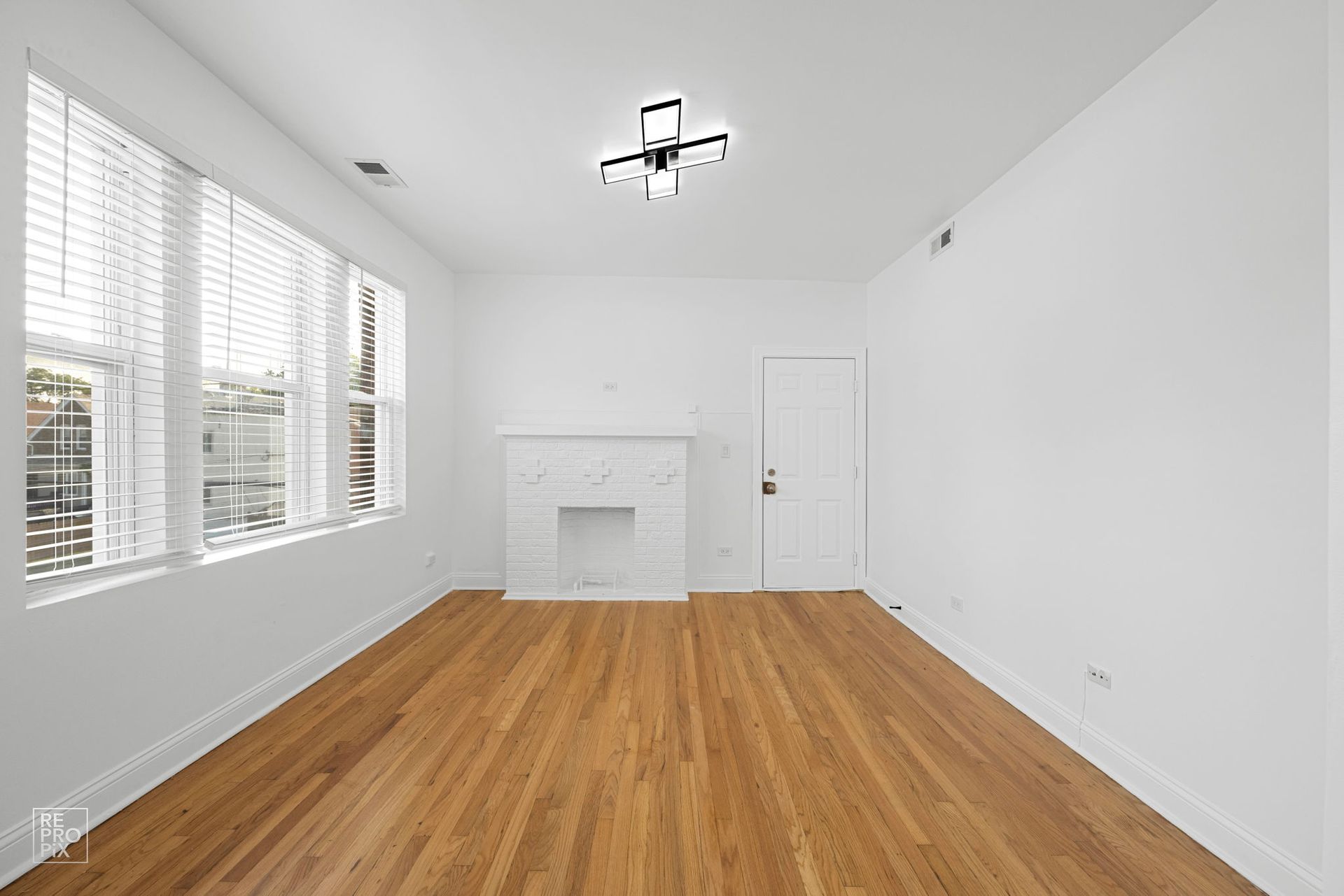 Empty room with wood floors, white walls, fireplace, windows with blinds, and a light fixture.