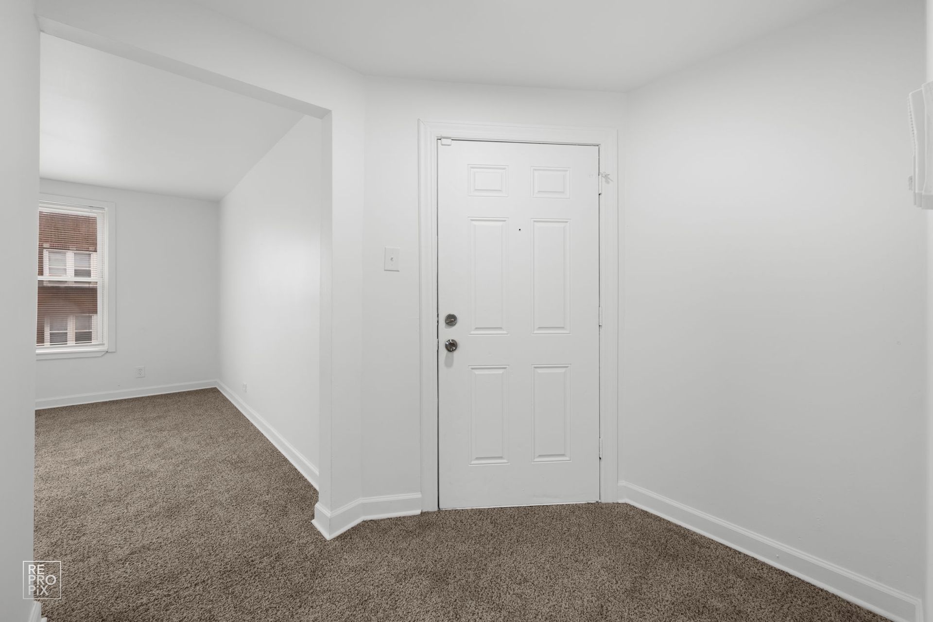 White-walled interior with a closed door. Brown carpet in the foreground and visible in the adjacent room with a window.