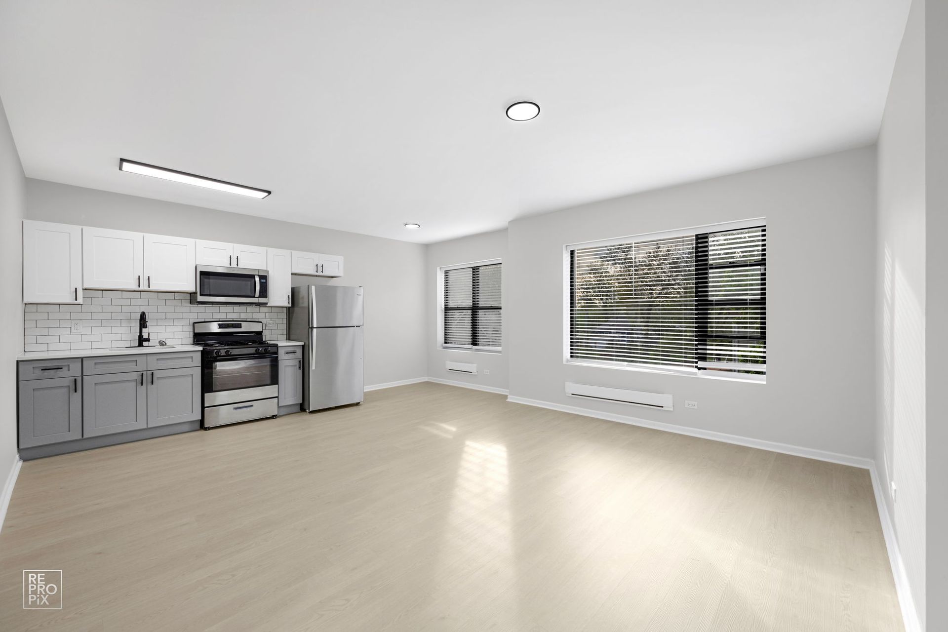 Empty, bright apartment interior with a kitchen, stainless steel appliances, and large windows.