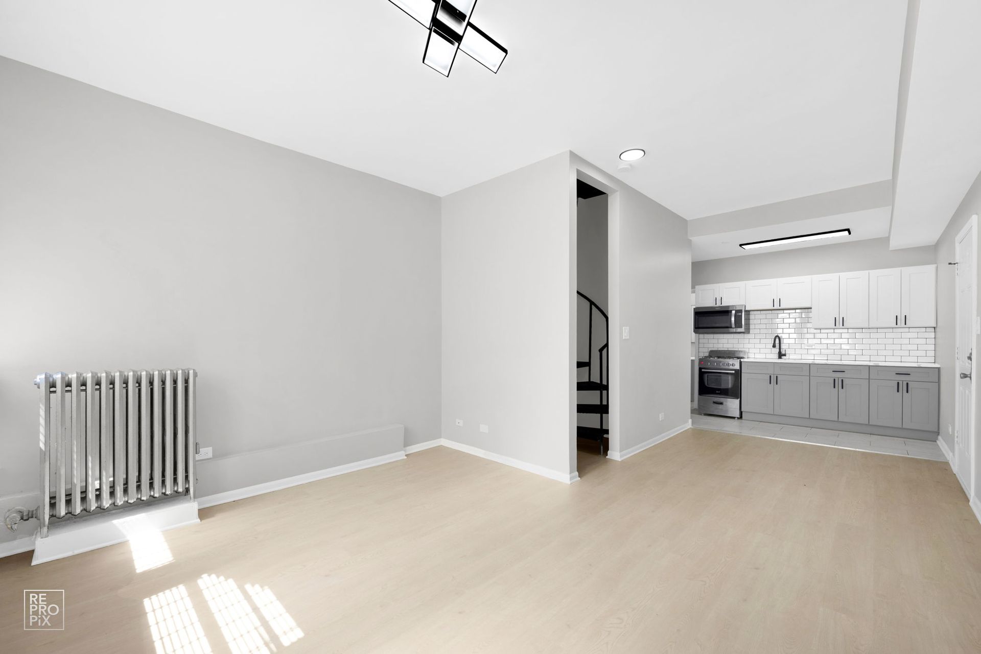 Empty apartment interior with light gray walls, light wood floor, kitchen, and spiral staircase.