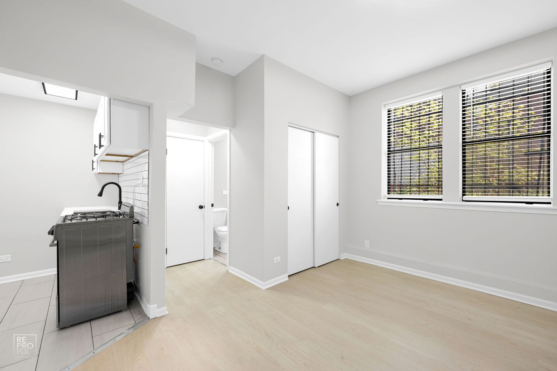 Small, empty apartment with a kitchenette, two windows, and a closet. Light gray walls, light wood floors.