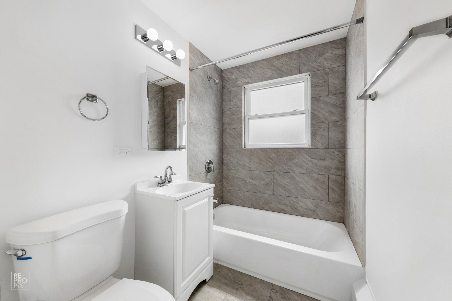 Small bathroom with white fixtures, gray tiled shower, and a small window.