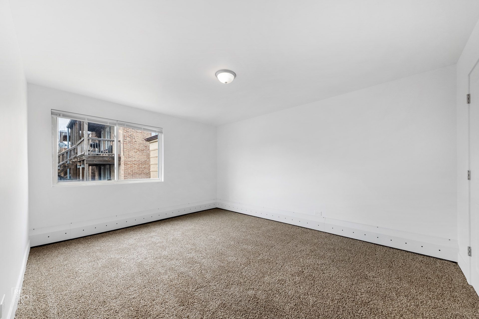 Empty room with neutral brown carpet, white walls, small window, and a single ceiling light.