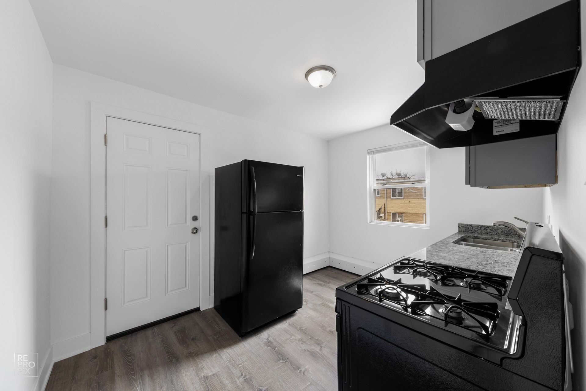Black appliances in a small, white kitchen: refrigerator, stove, exhaust hood, and a door.