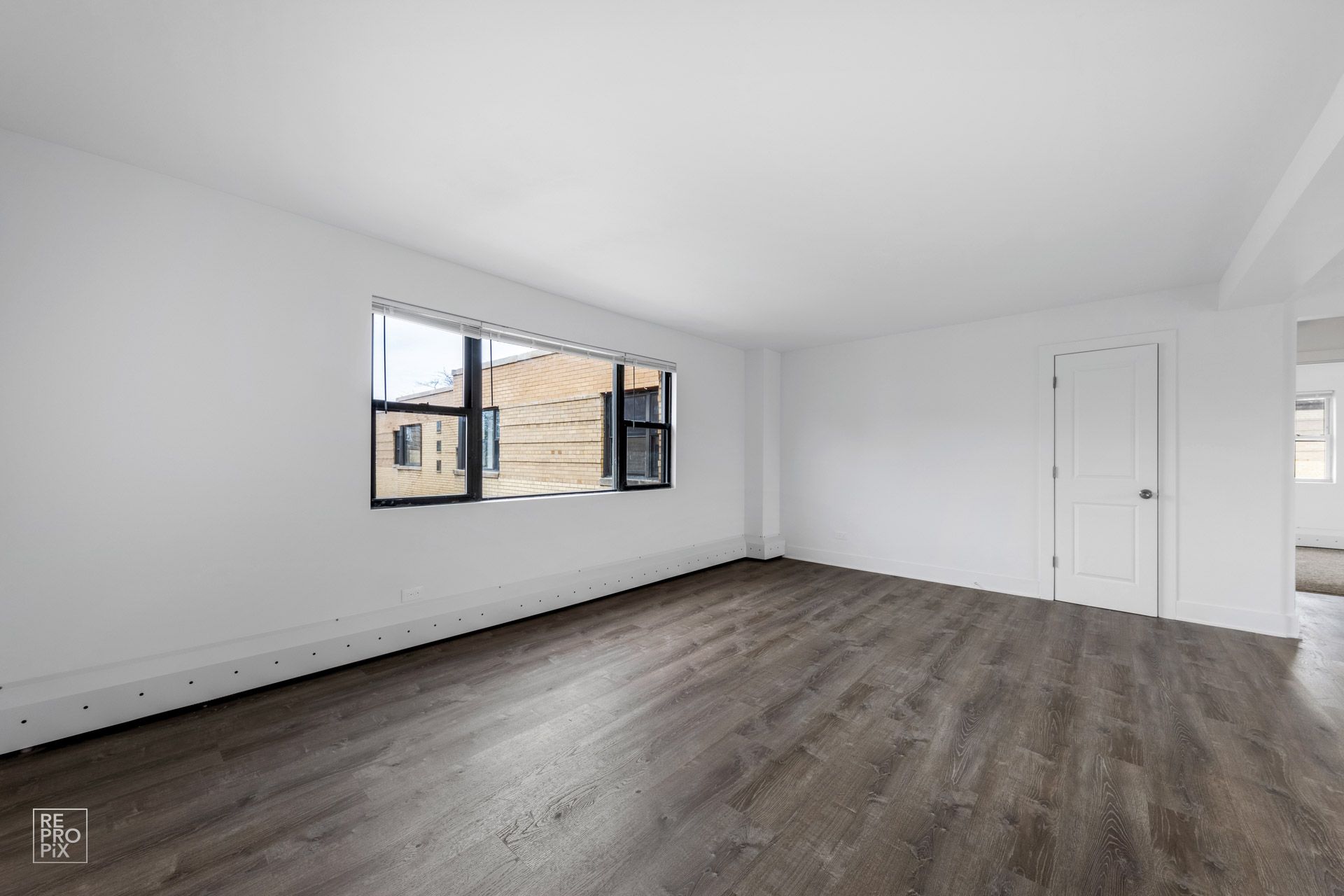 Empty room with a large window, white walls, dark wood-look floor, and a closed white door.