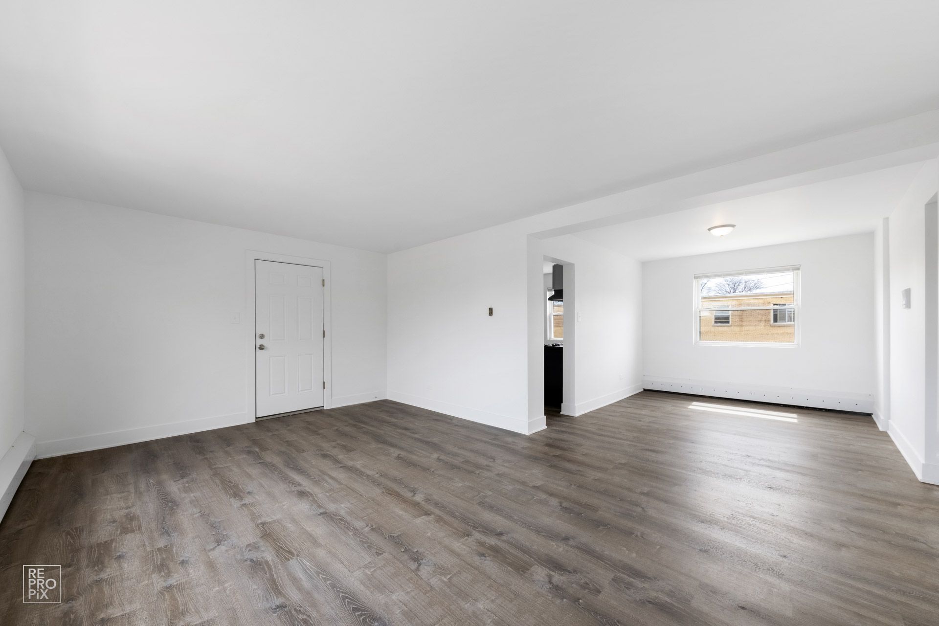 Empty living room with wood-look flooring, white walls, and a doorway to a kitchen.