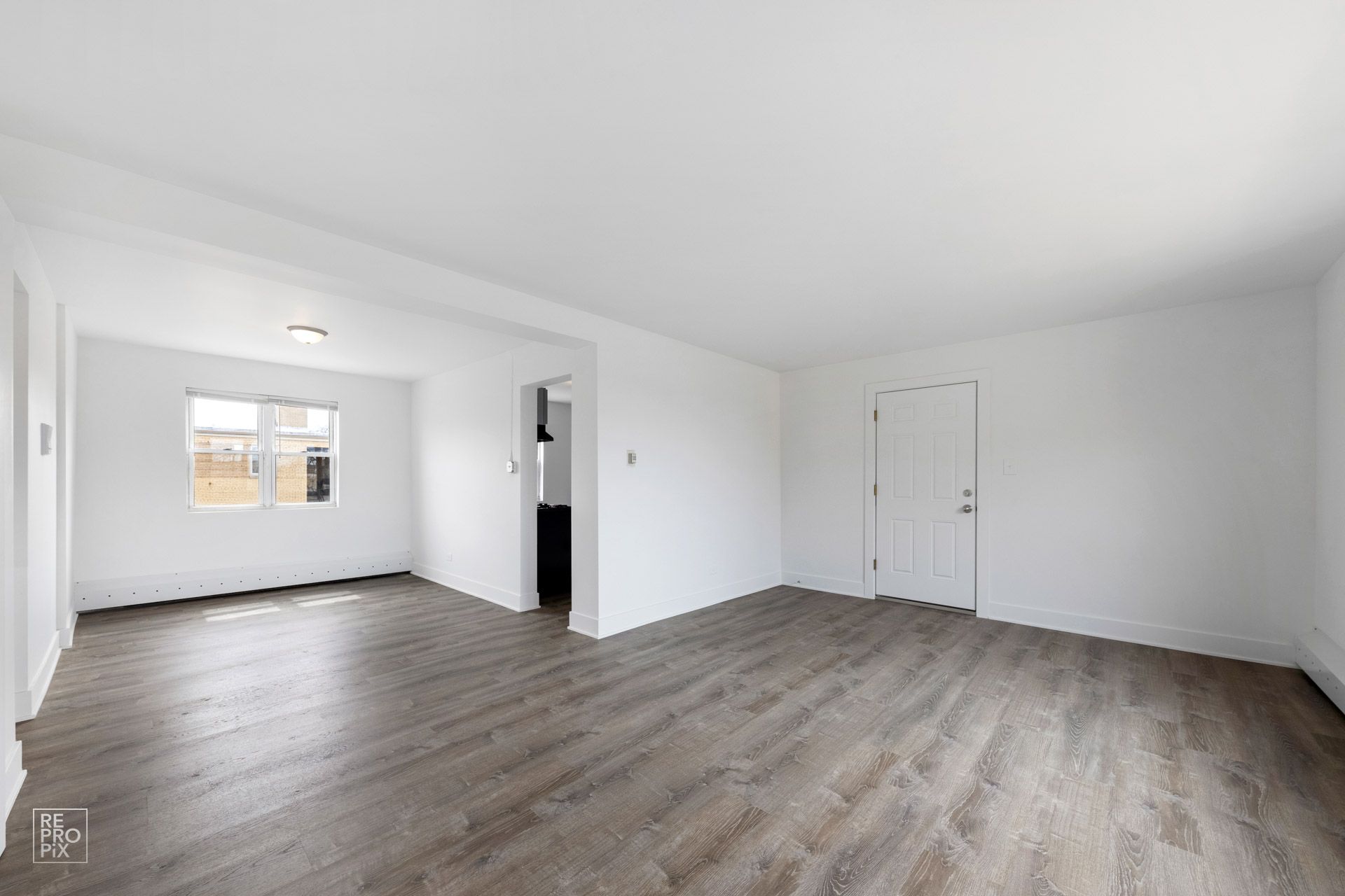 Empty room with wood-look flooring, white walls, a window, and a door; a glimpse of a dark kitchen area.