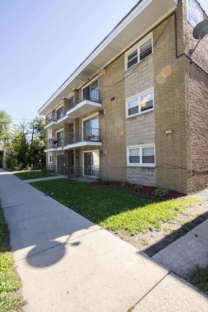 Three-story brick apartment building with balconies, sidewalk, and green lawn on a sunny day.
