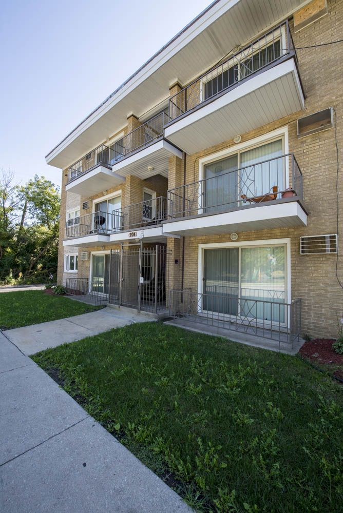 Brick apartment building with balconies, sidewalk, and green lawn on a sunny day.