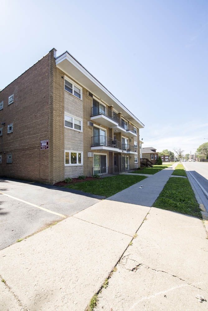 Three-story brick apartment building with balconies, sidewalk, and parking on a sunny day.