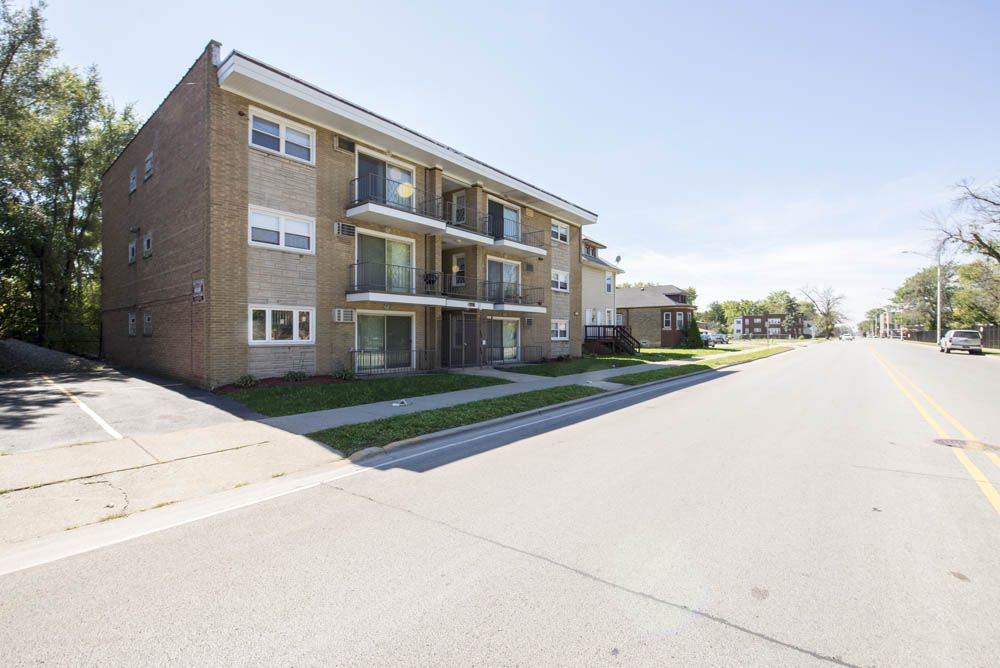 Brick apartment building on a street, with balconies and parking spaces. Sunny day.